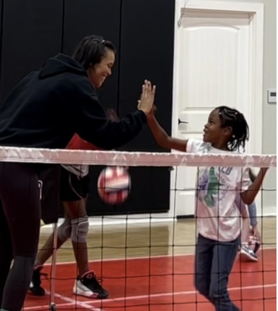 Adult and child high-five across a volleyball net indoors, with a door in the background.