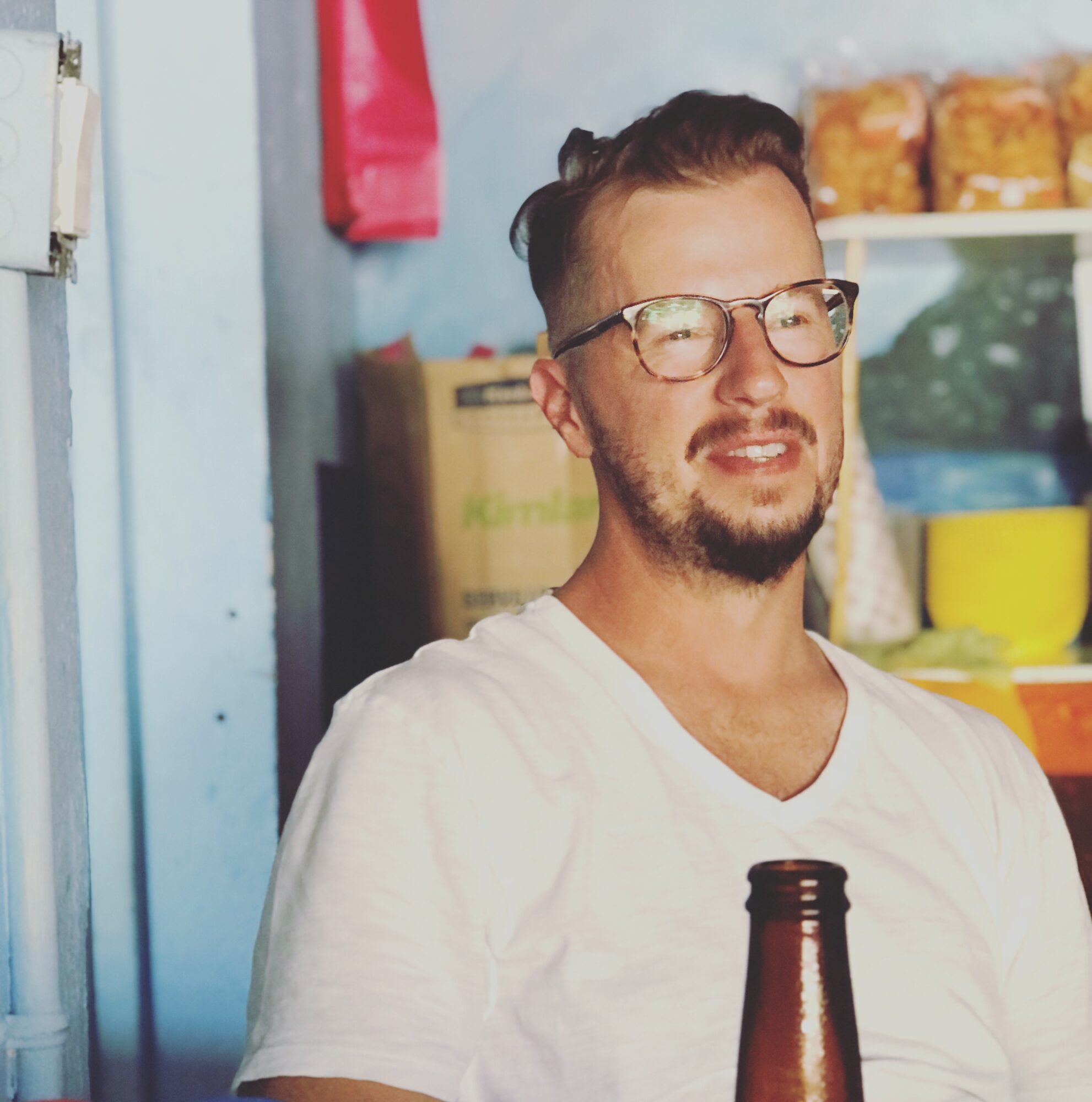 Man with glasses and beard smiling, wearing a white shirt, in a store with shelves of snacks behind him.
