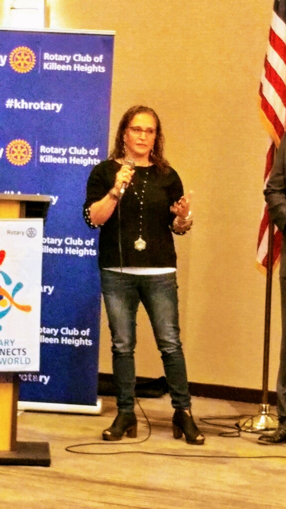 Woman speaking into microphone on stage with Rotary Club banners and American flag behind her.