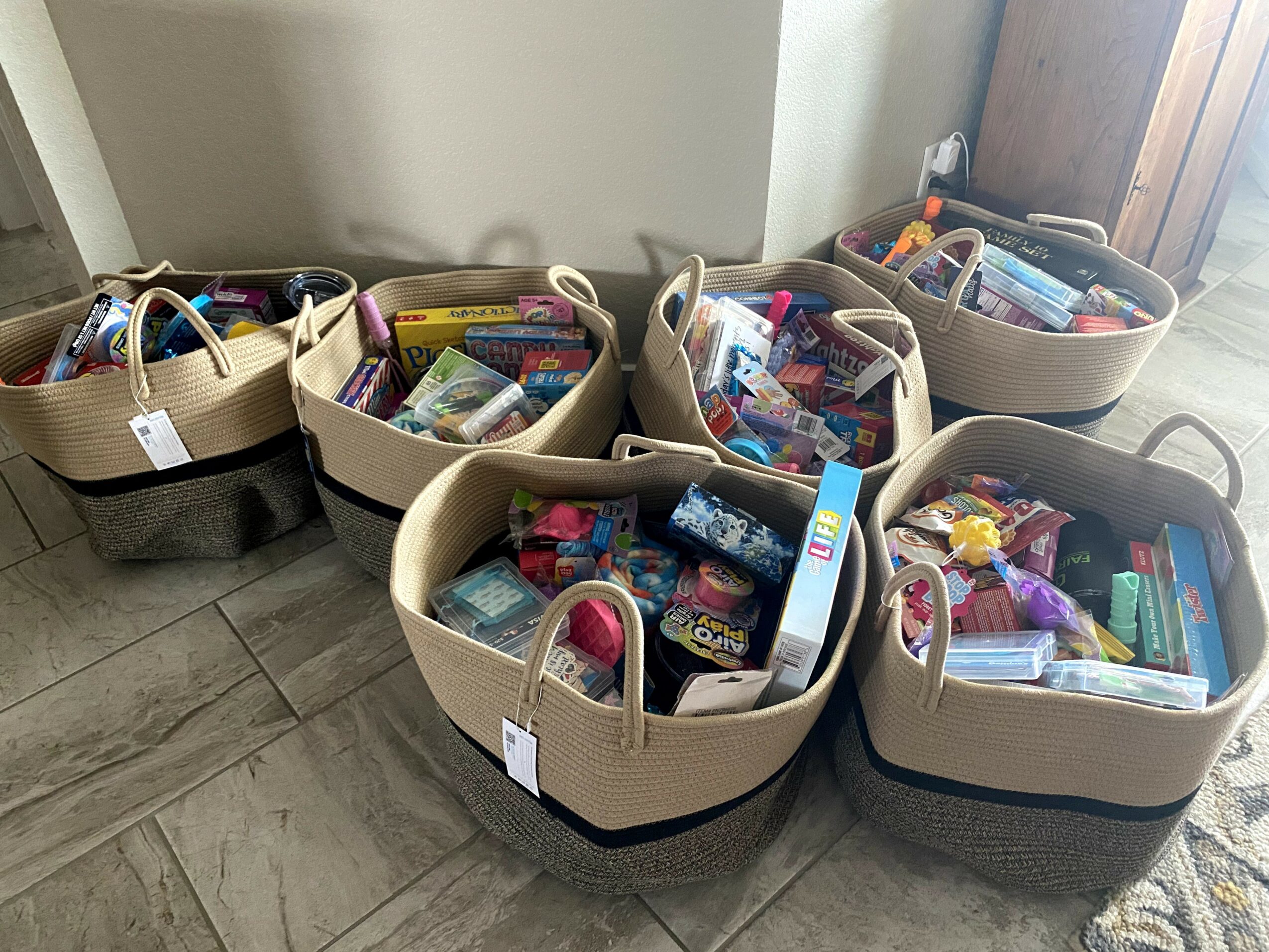 Five beige baskets filled with snacks and food items are placed on a wooden floor near a wall and door.