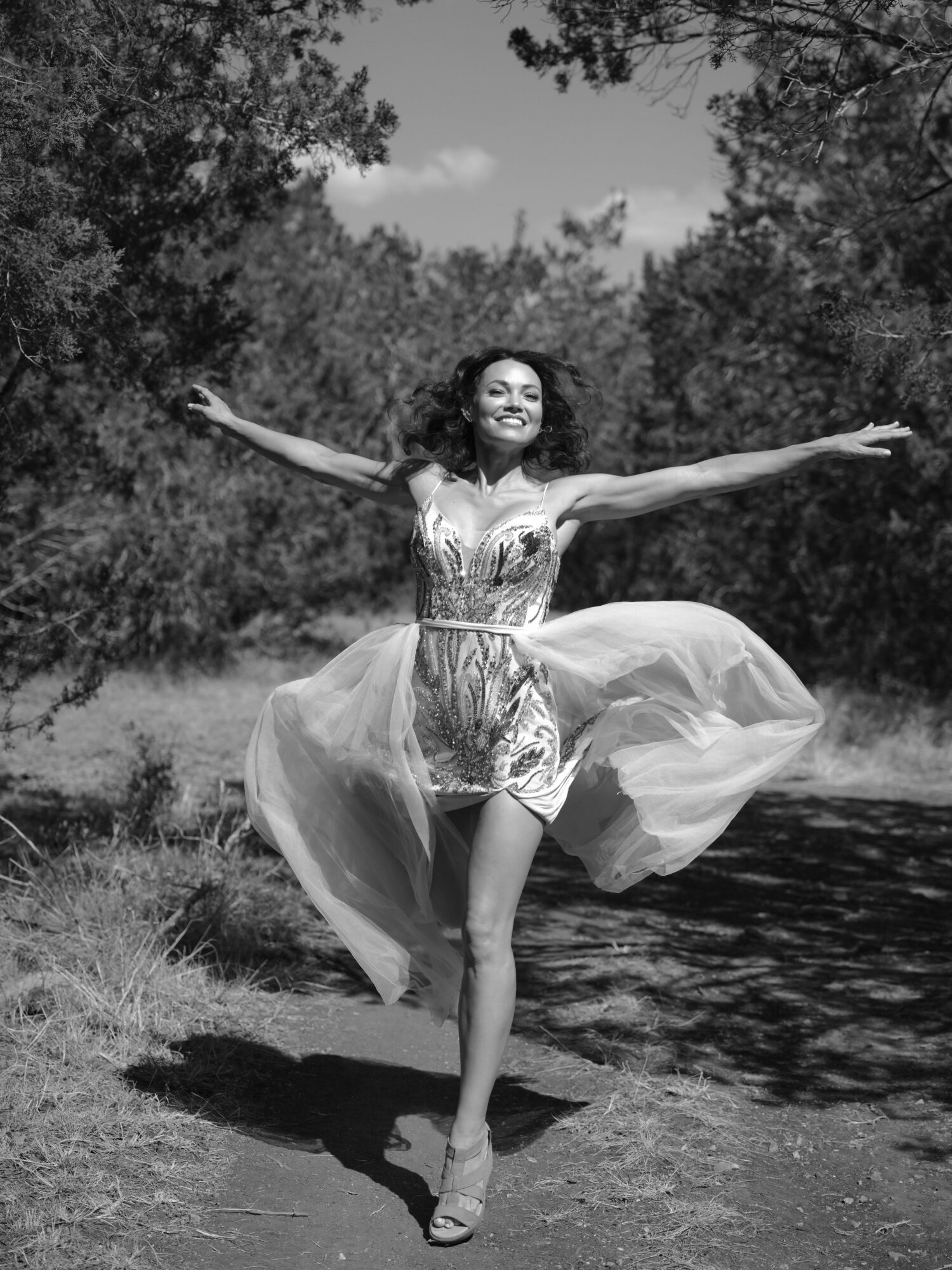 Woman in dress dancing outdoors near water with arms outstretched and smiling.