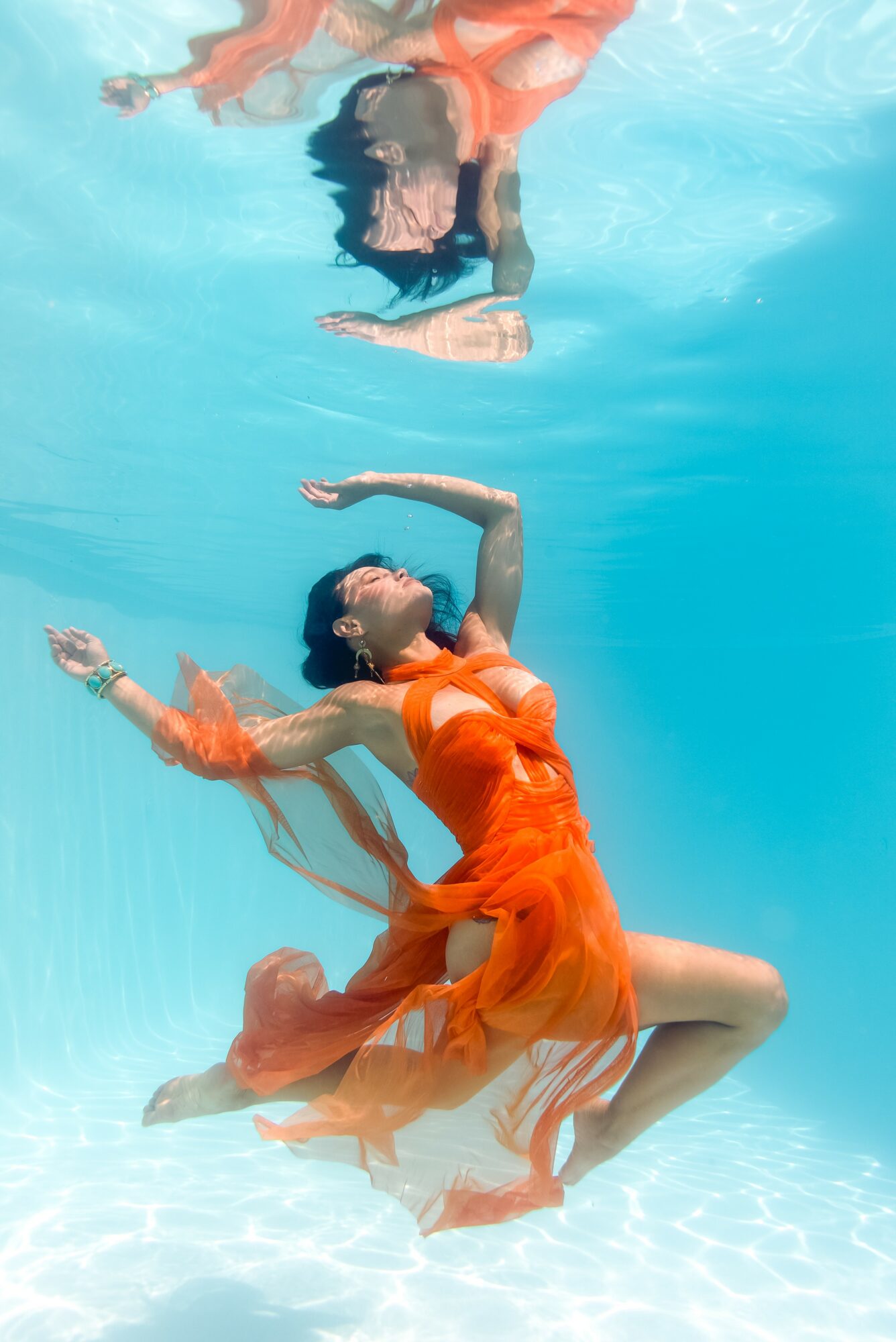 Woman in orange dress floating underwater with arms extended and eyes closed.