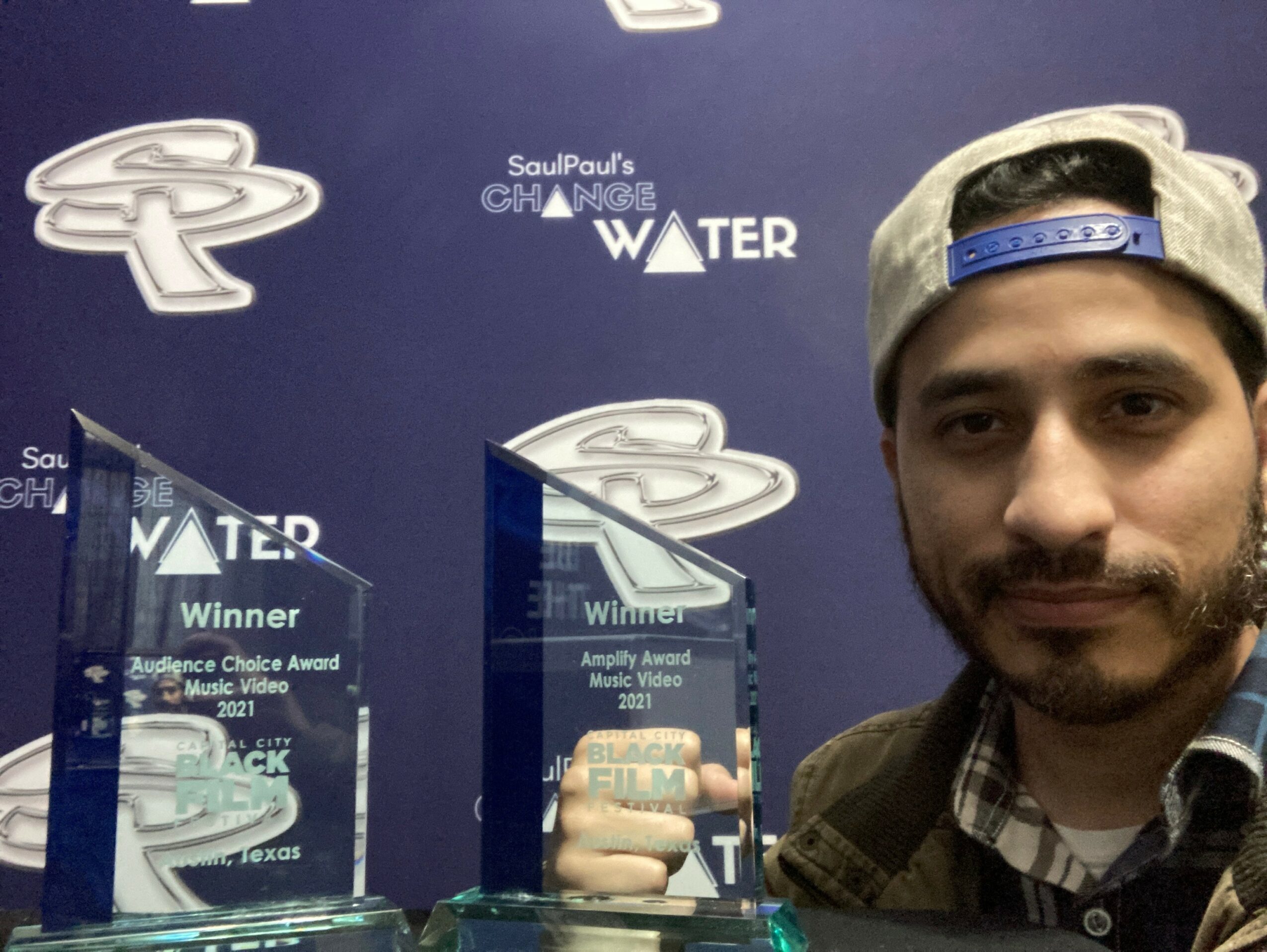 Man wearing a cap and plaid shirt taking a selfie in front of award display with blue background.