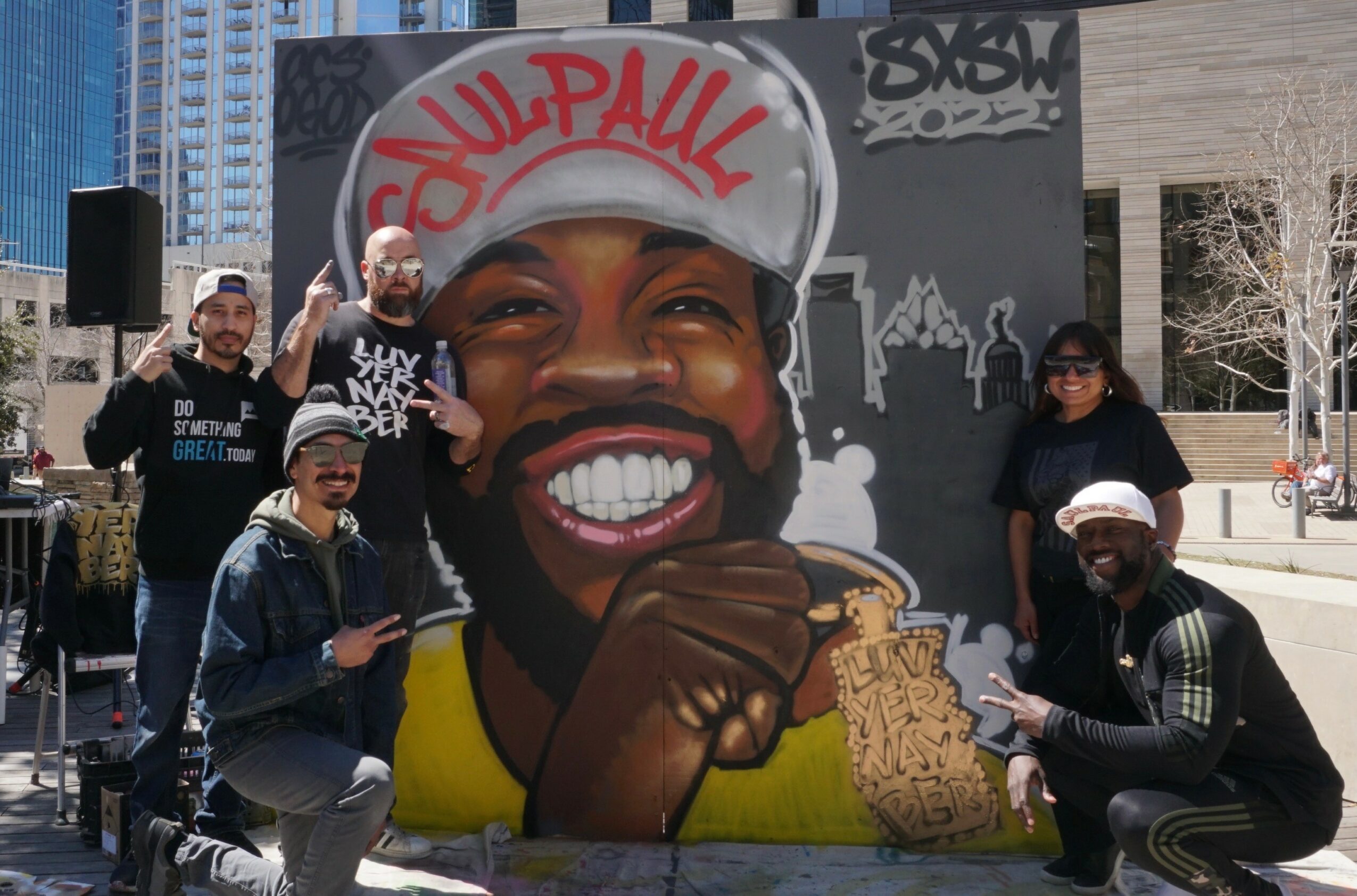 Group of five people posing in front of a large mural of a smiling man wearing a white cap with red writing, holding a sandwich.