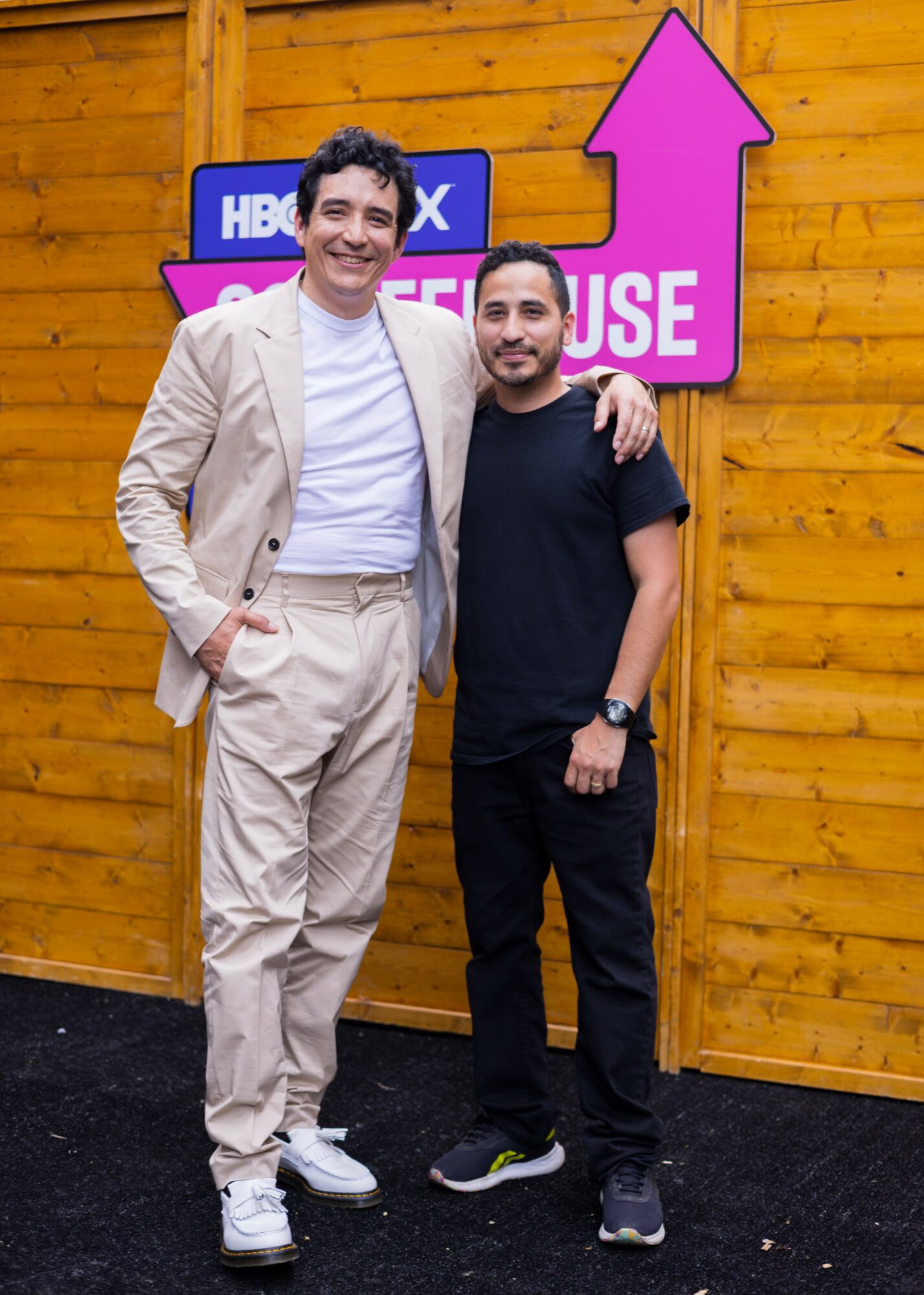 Two men standing side by side, smiling, with a wooden wall and purple arrow sign behind them.