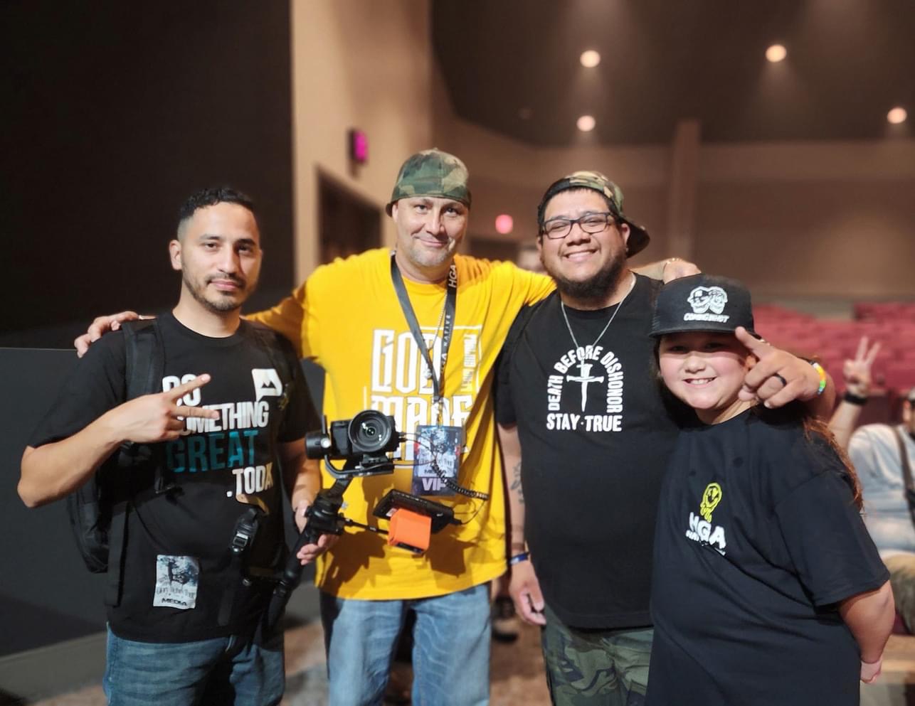 Four people standing together indoors, smiling, with one holding a camera, in a dimly lit room.