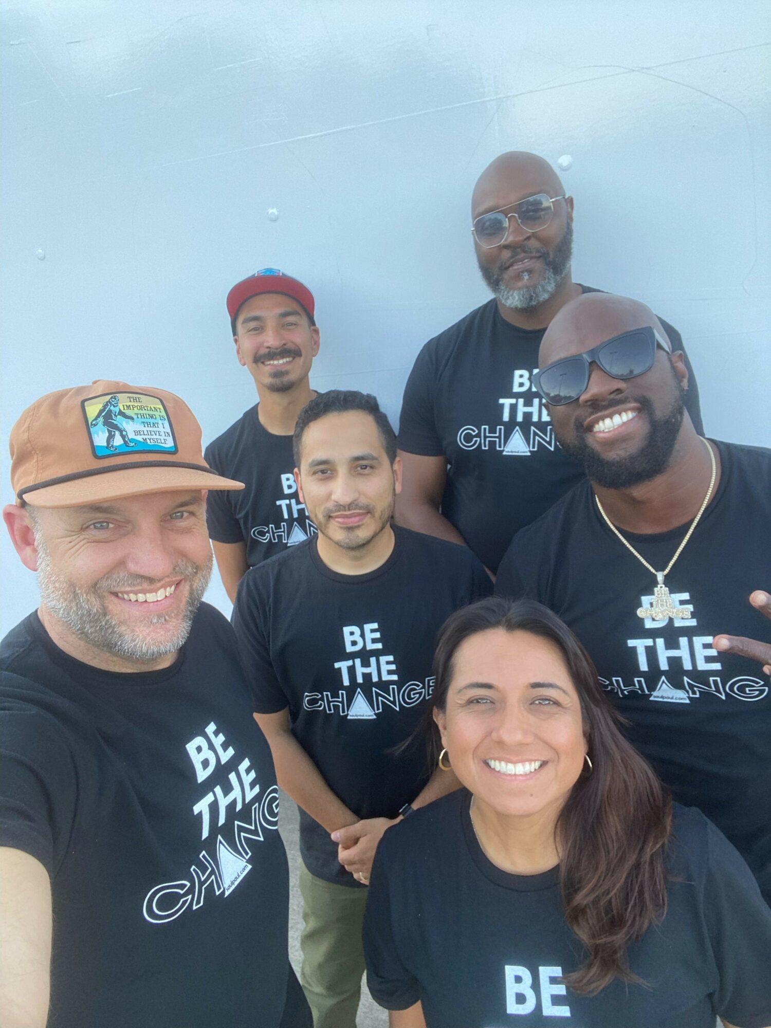 Group of six people smiling outdoors, wearing matching black t-shirts with text, some wearing sunglasses and hats.