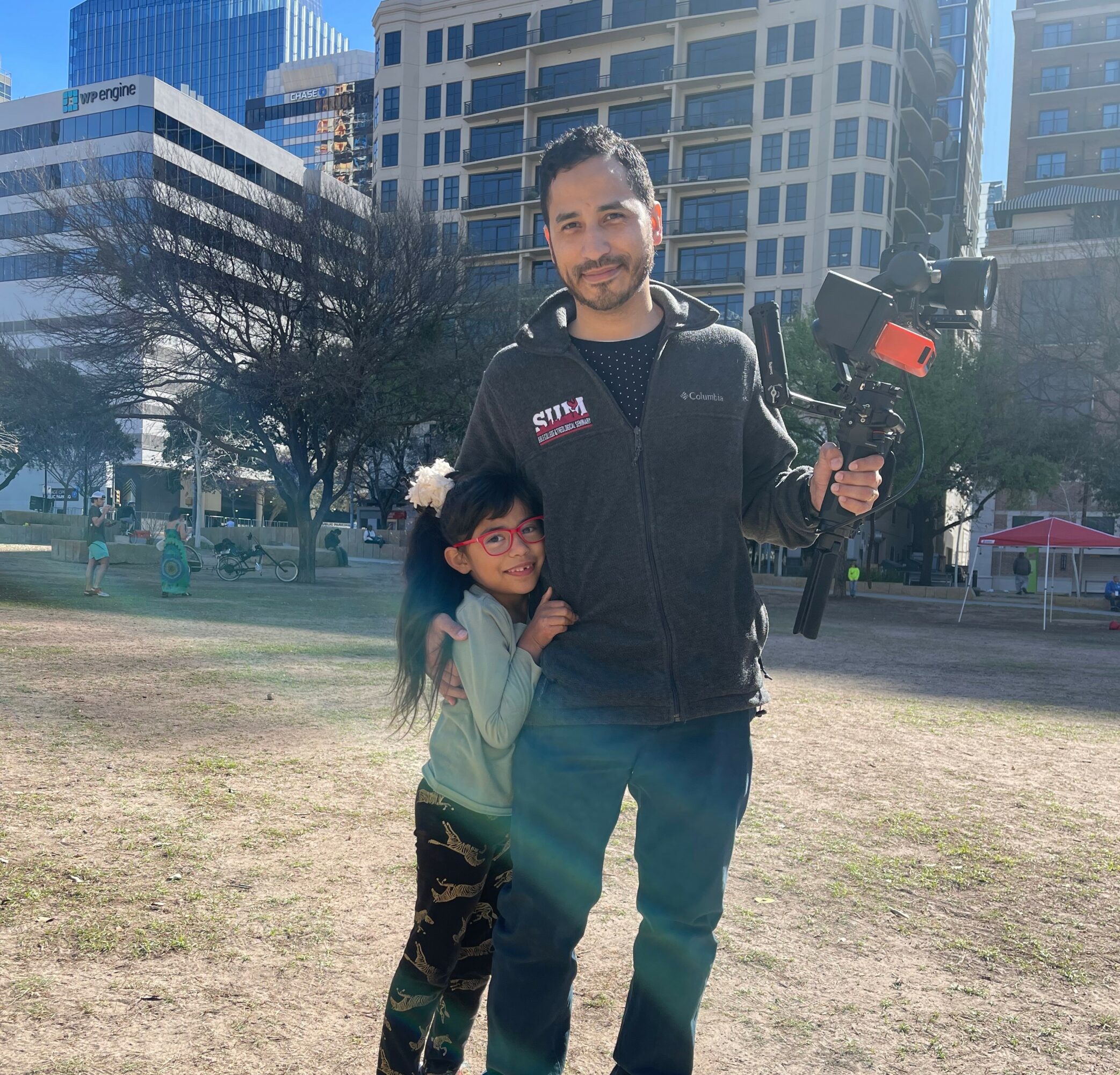 Man holding a camera with a young girl standing beside him outdoors in a city park.