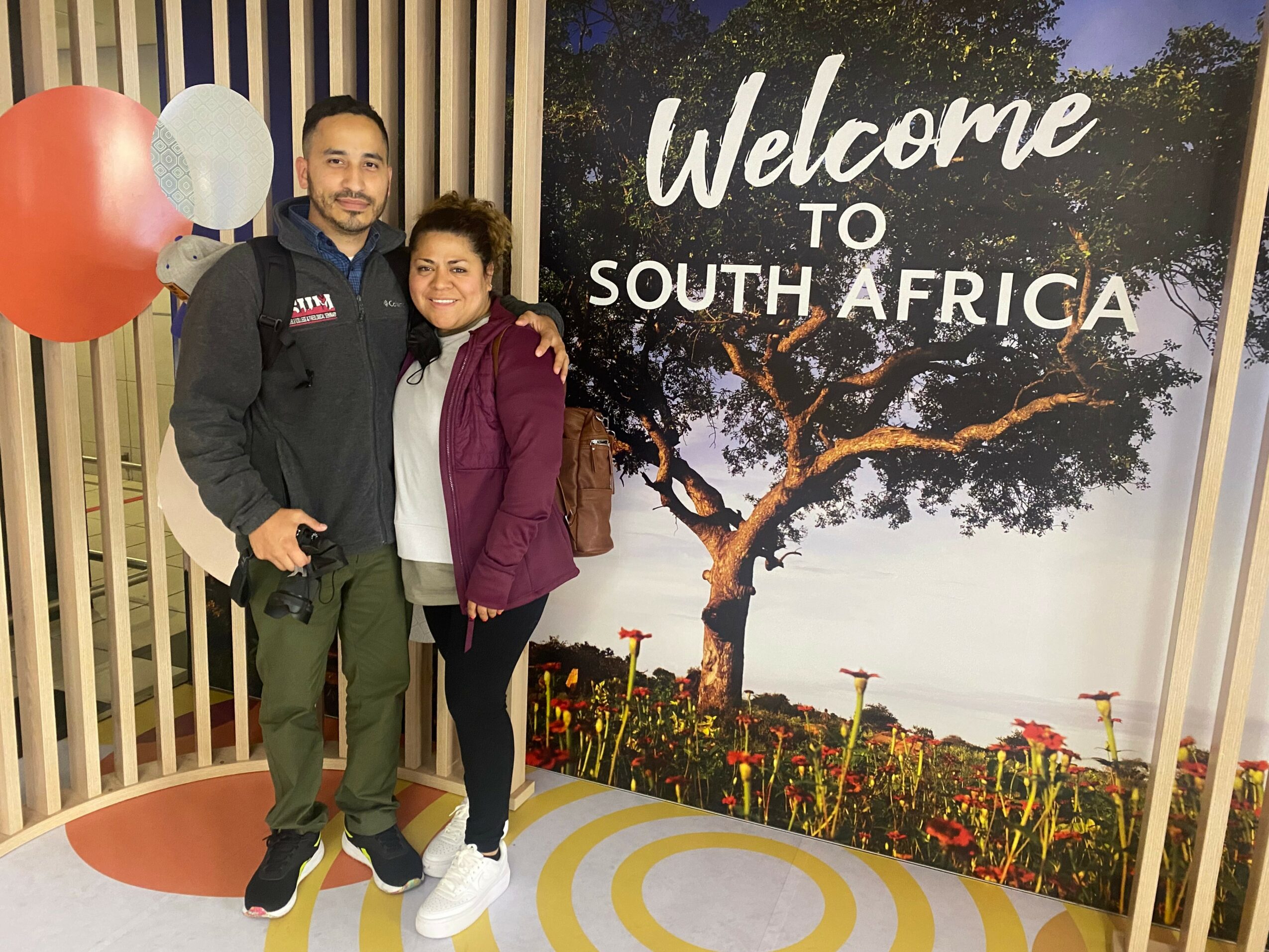 Two people standing in front of a South Africa welcome sign, smiling, with balloons nearby.