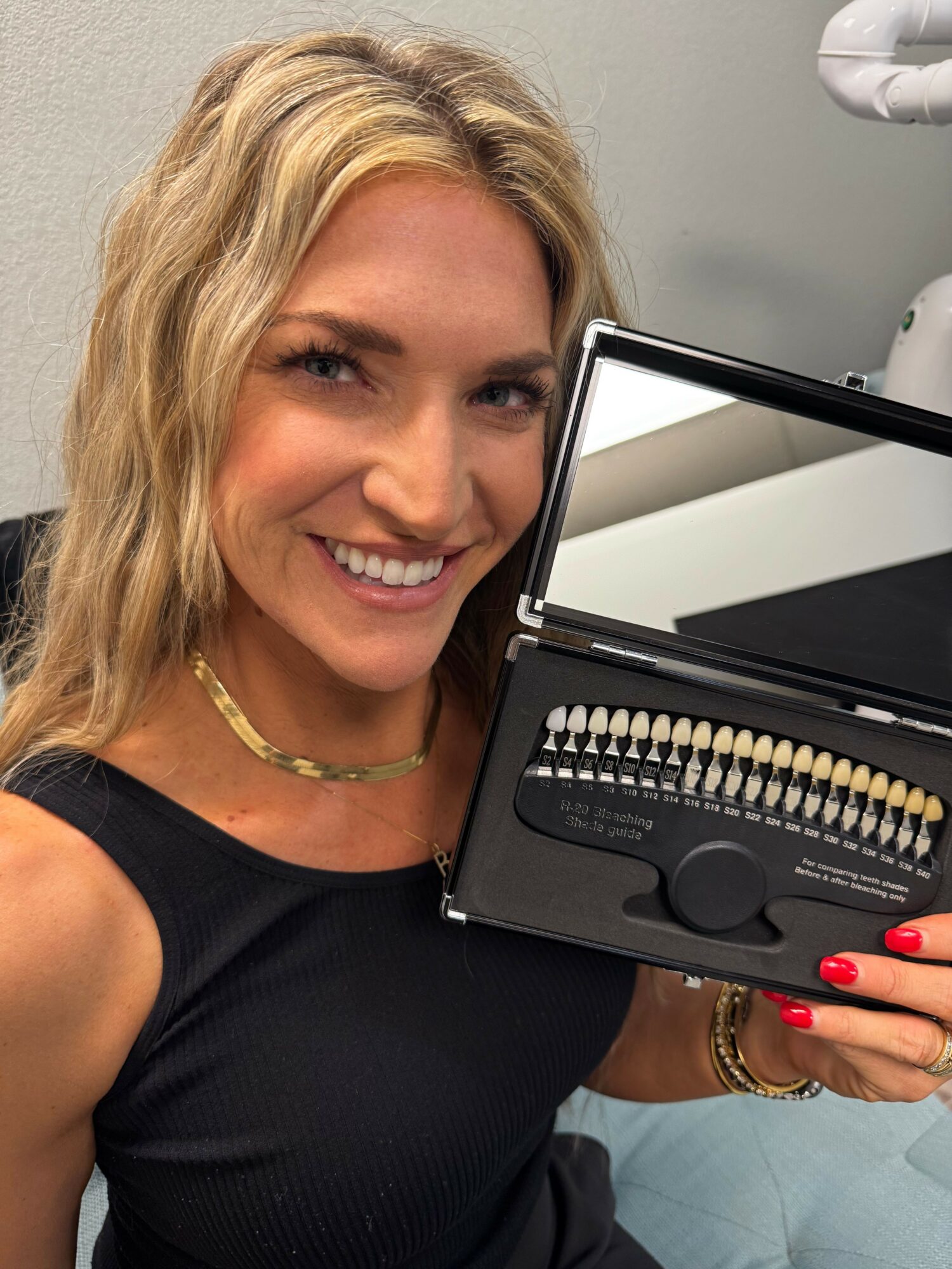 Woman with blonde hair smiling, holding a dental shade guide in a case, wearing a black top and gold jewelry.