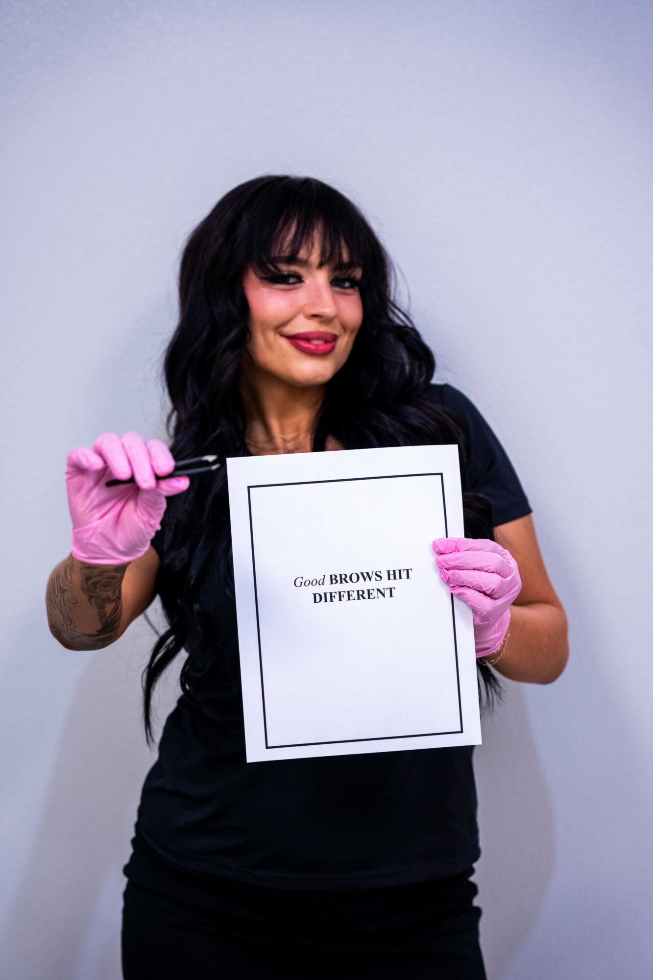 Woman with dark hair and pink gloves holding a sign with text, smiling, against a plain background.