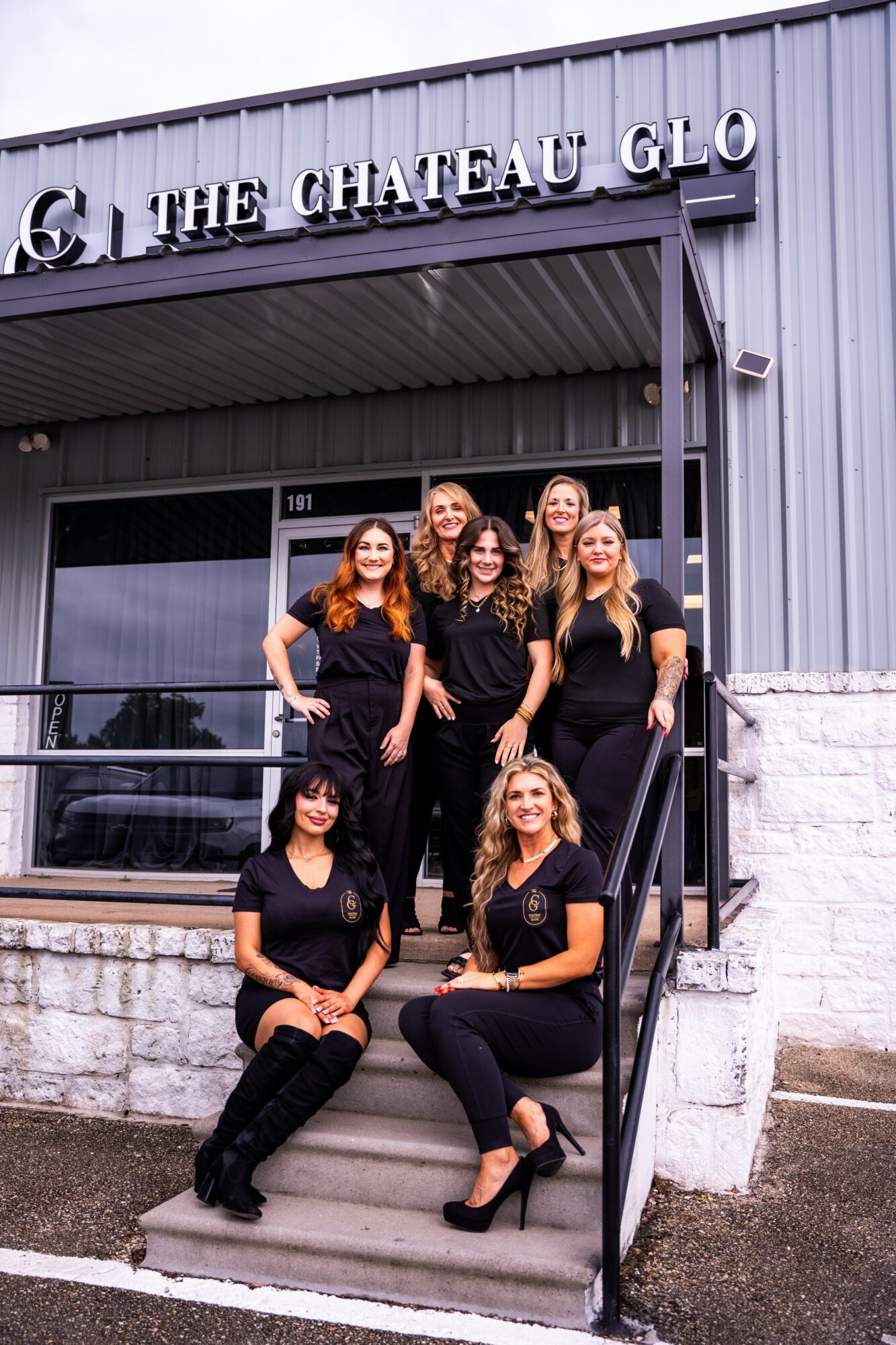 Six women in black outfits pose on steps outside a building with a sign reading 'The Chateau Glo'.