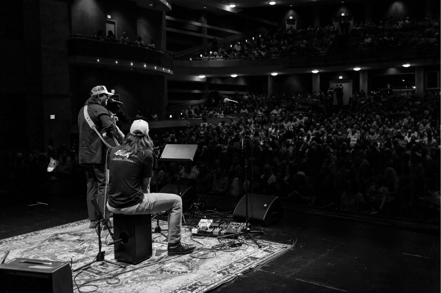 Two musicians perform on stage with a large audience in the background, one playing guitar and the other singing.