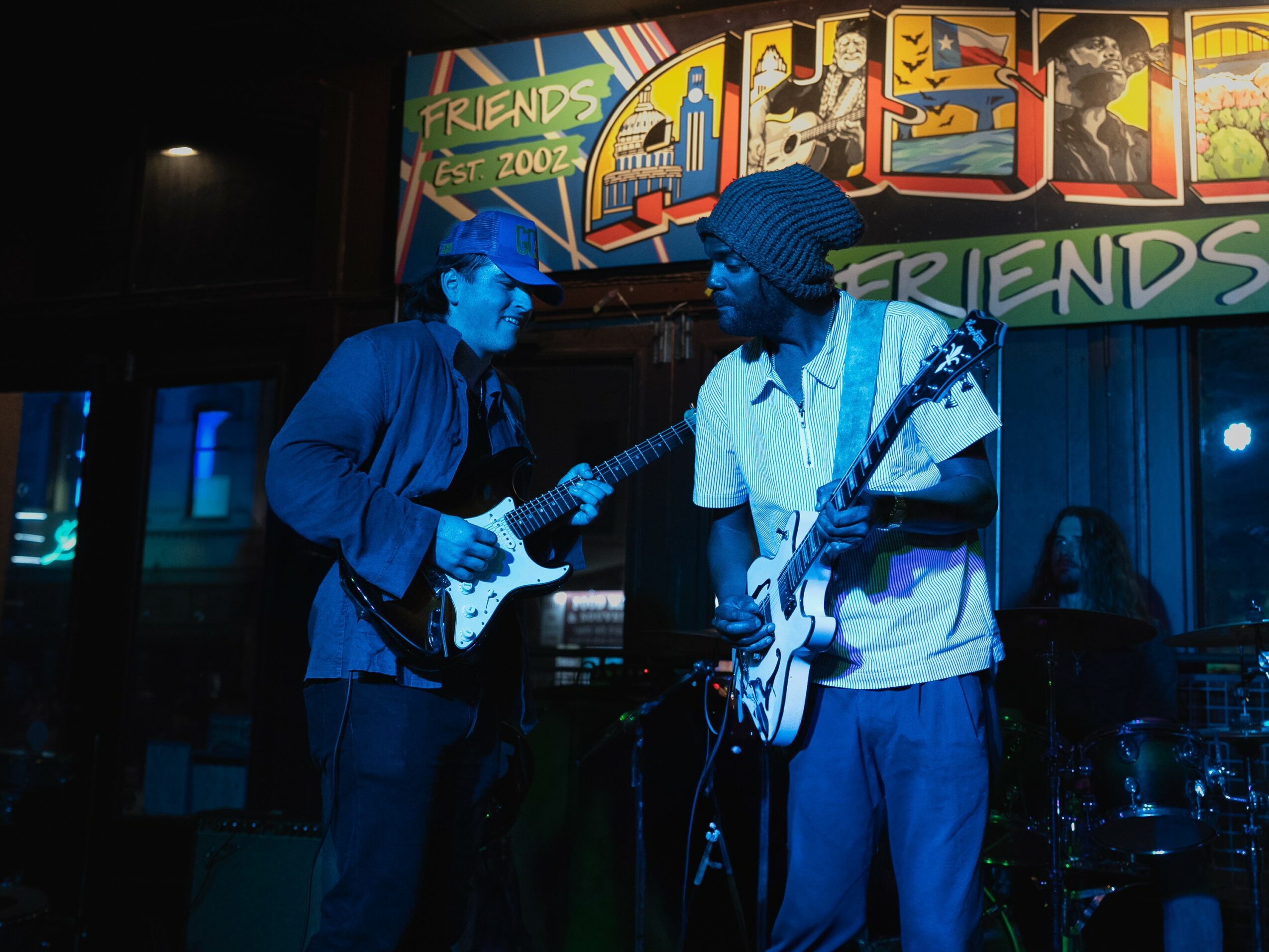 Two musicians playing guitars on stage with colorful banners and a dark background.