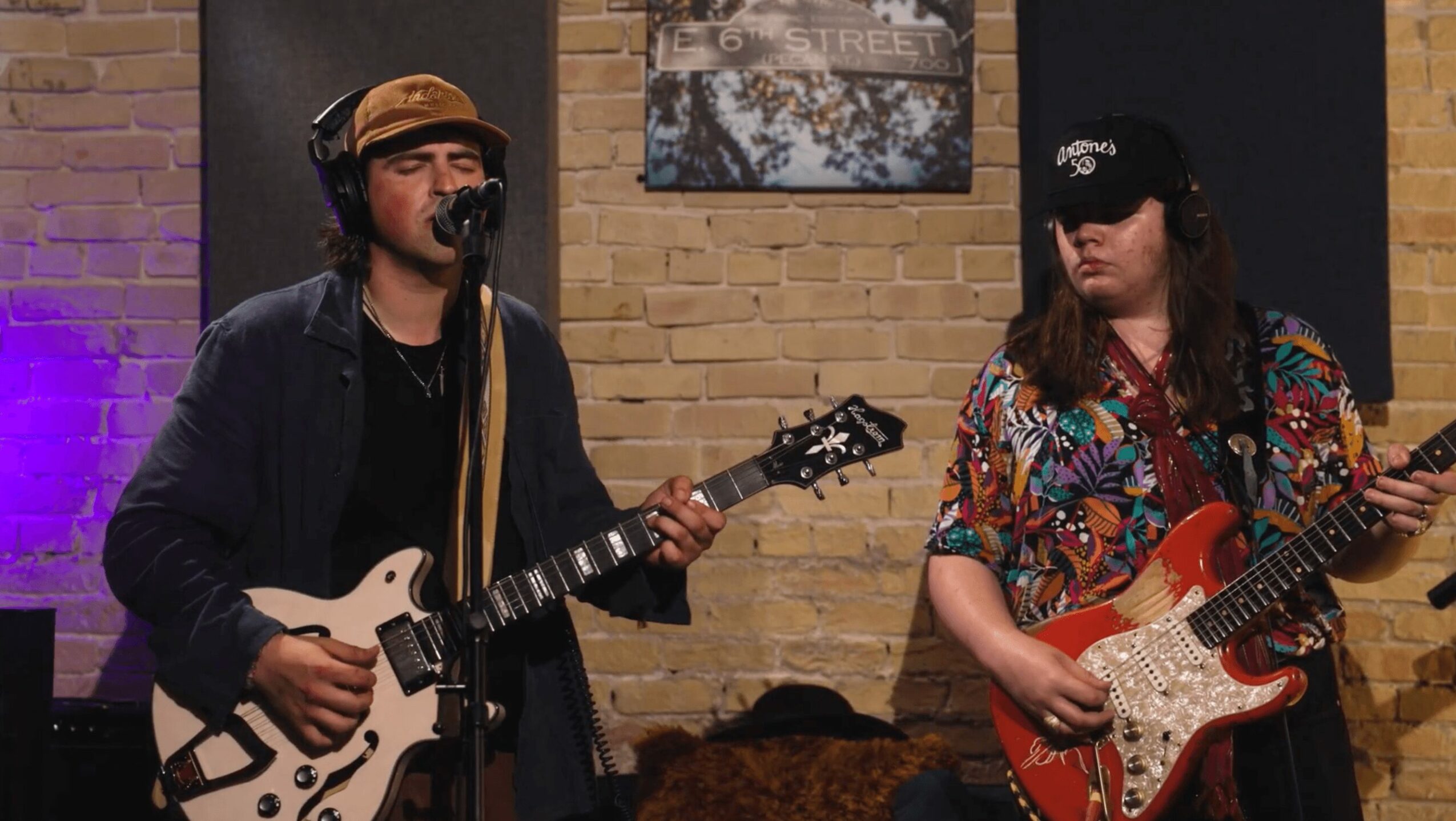 Two musicians playing guitars in a brick-walled room with artwork and purple lighting.