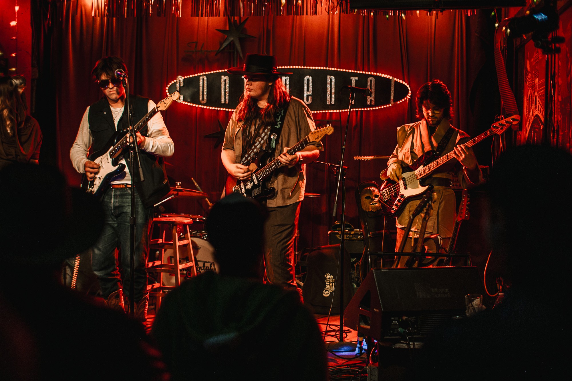 Four musicians perform on stage with guitars, microphone, and drums, in a dimly lit venue with red curtains.