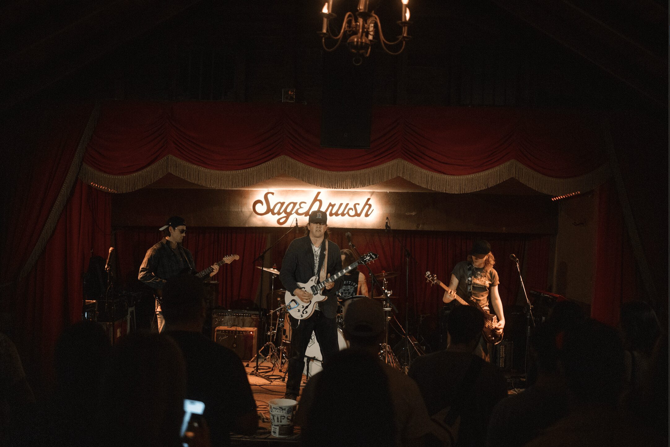 Band performs on stage with red curtains and a chandelier, with a sign reading 'Sagebrush' behind them.