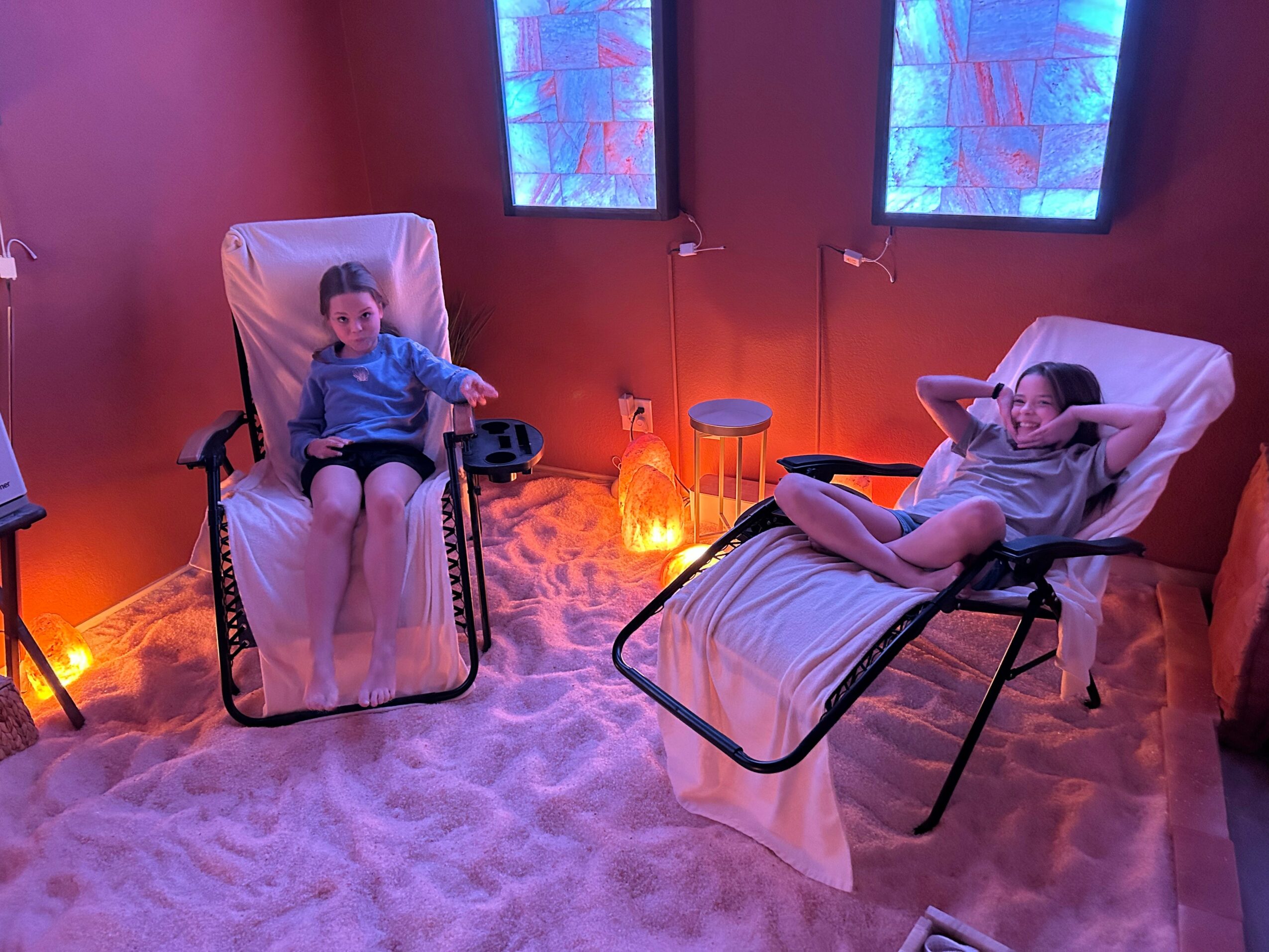 Two women relax on lounge chairs in a room with pink walls and blue-lit windows, sandy floor, and warm lighting.