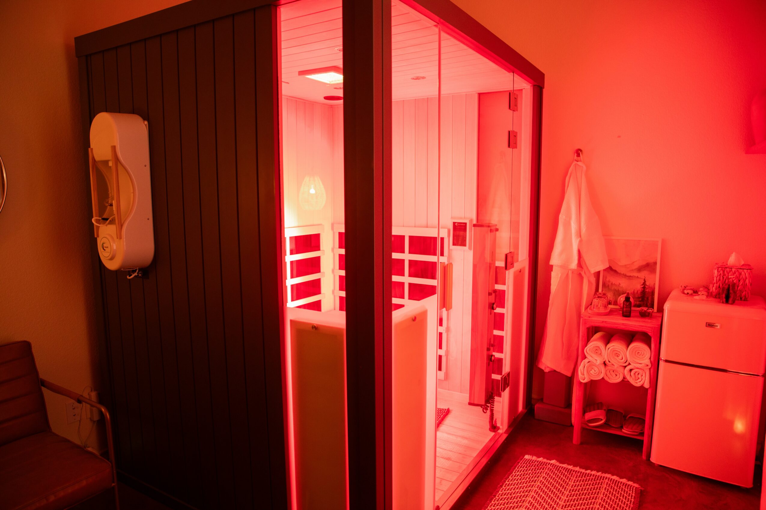 Small sauna room with glass door, red lighting, and wooden interior, next to a towel rack and small refrigerator.