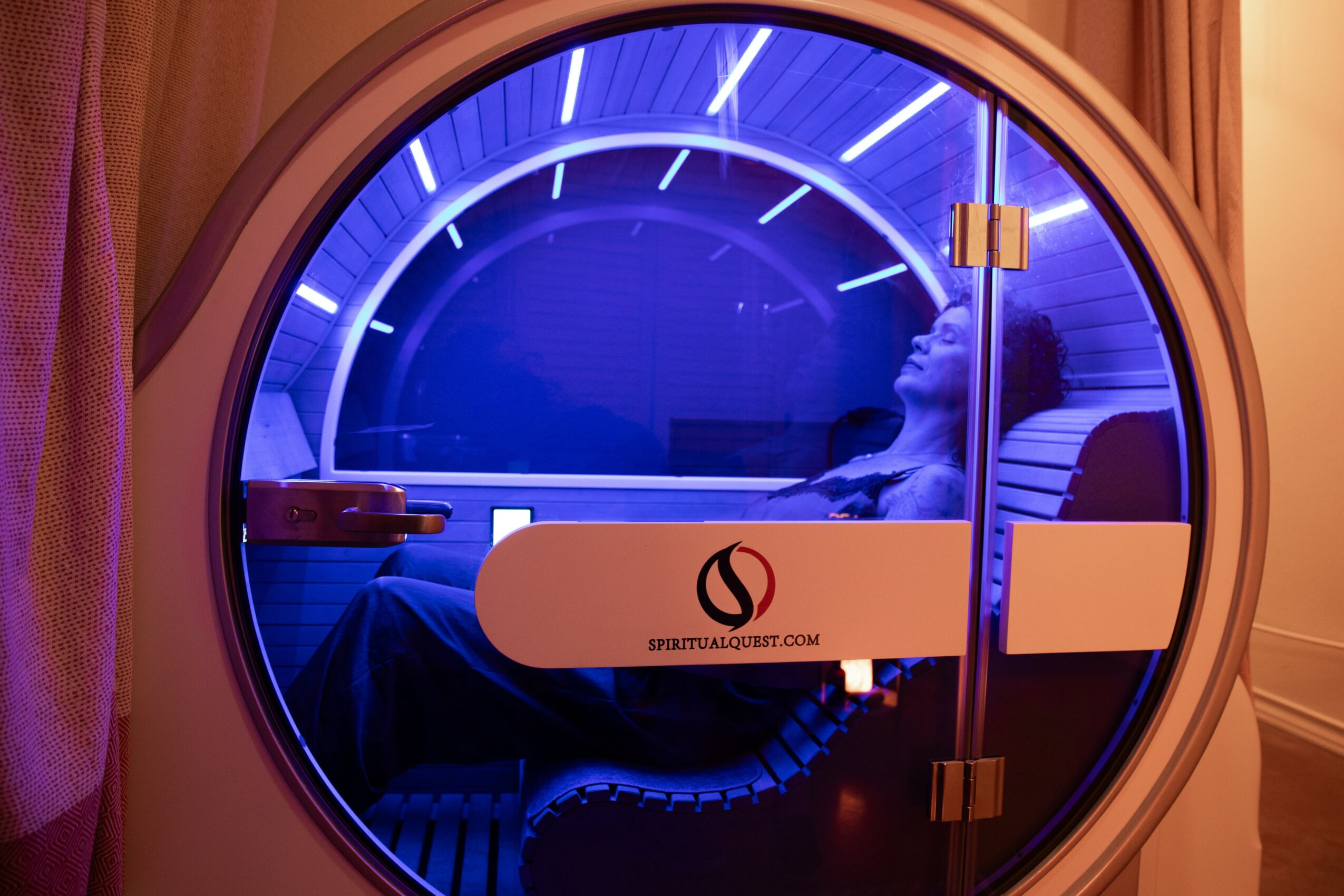 Person inside a circular hyperbaric chamber with blue lighting, sitting and looking to the side, with a logo on the chamber door.