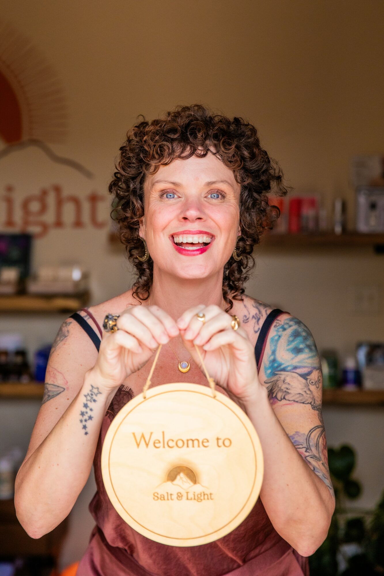 Woman with curly hair smiling, holding a round sign with text, in a room with shelves and decorations.