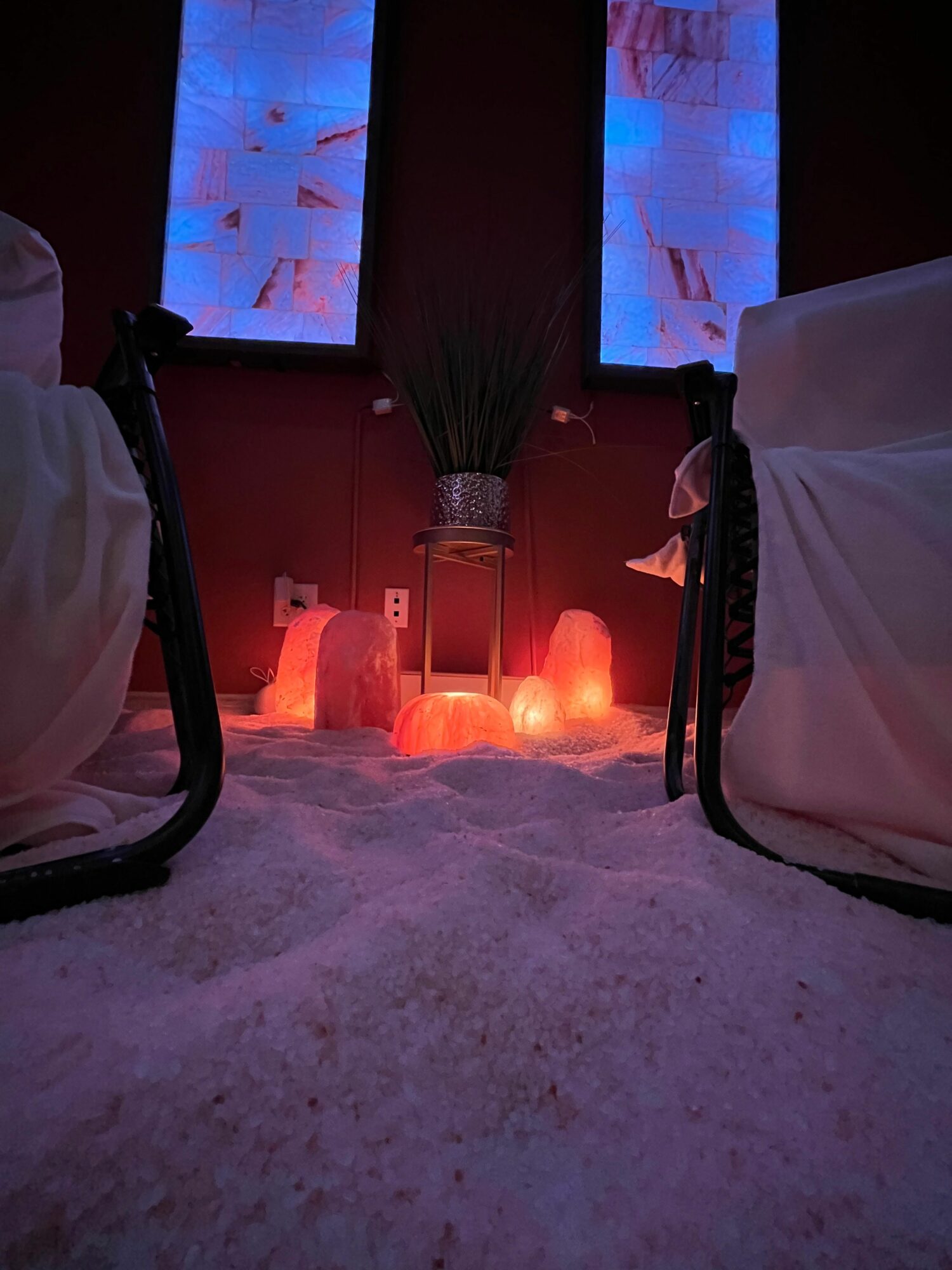 Cozy indoor scene with salt floor, salt lamps, potted plant, and two chairs with white covers, illuminated by blue and red lights.