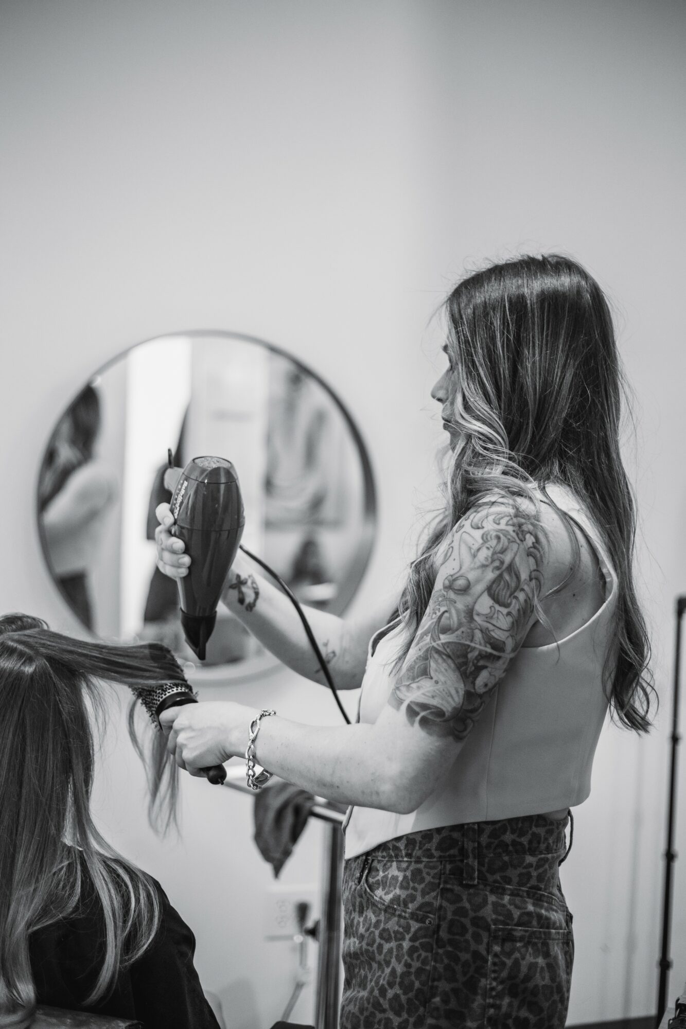 Woman styling hair in front of a mirror, holding a hairbrush, with another person seated nearby.