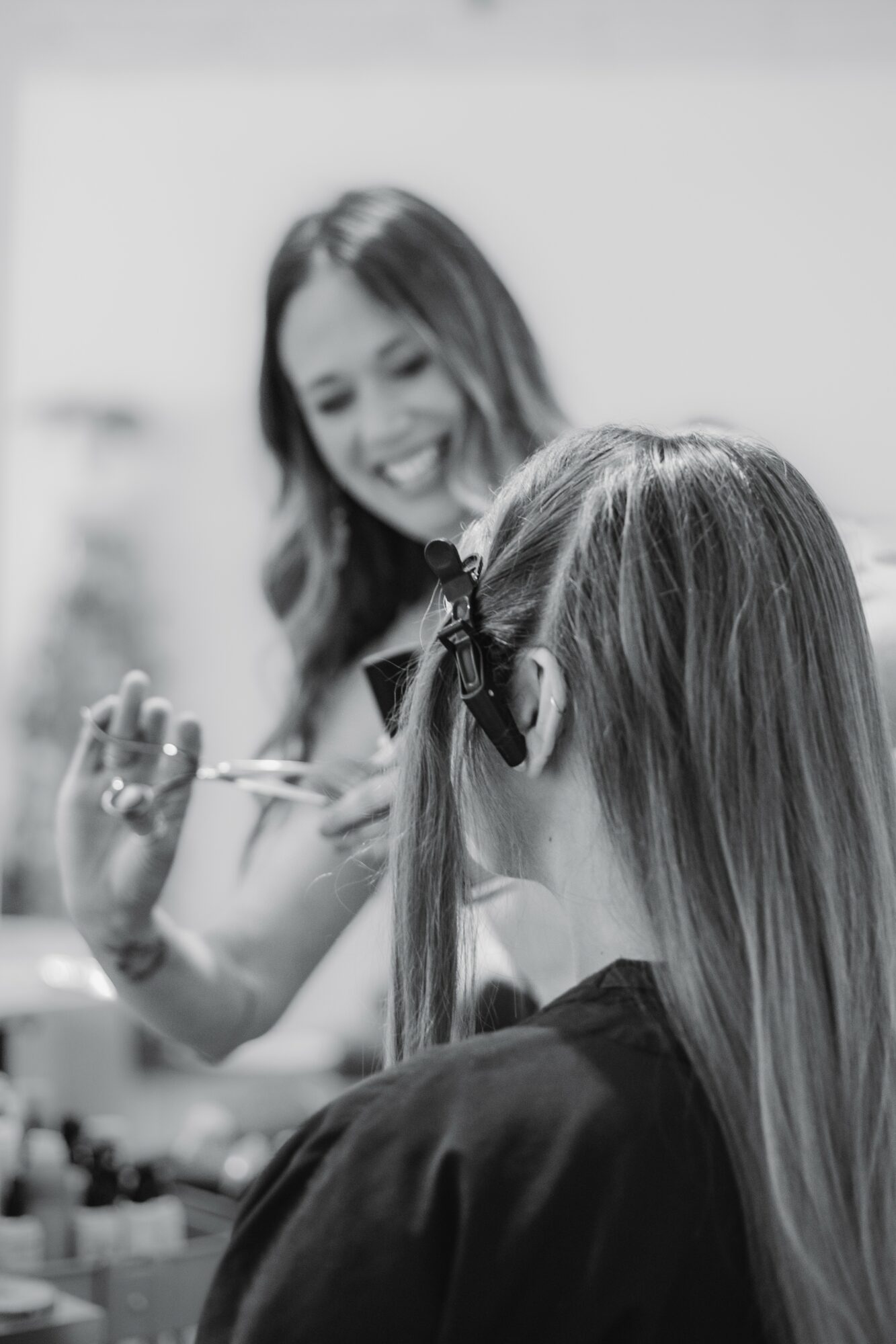 Two women smiling, one applying makeup with a brush, the other with long hair tied back with a bow.