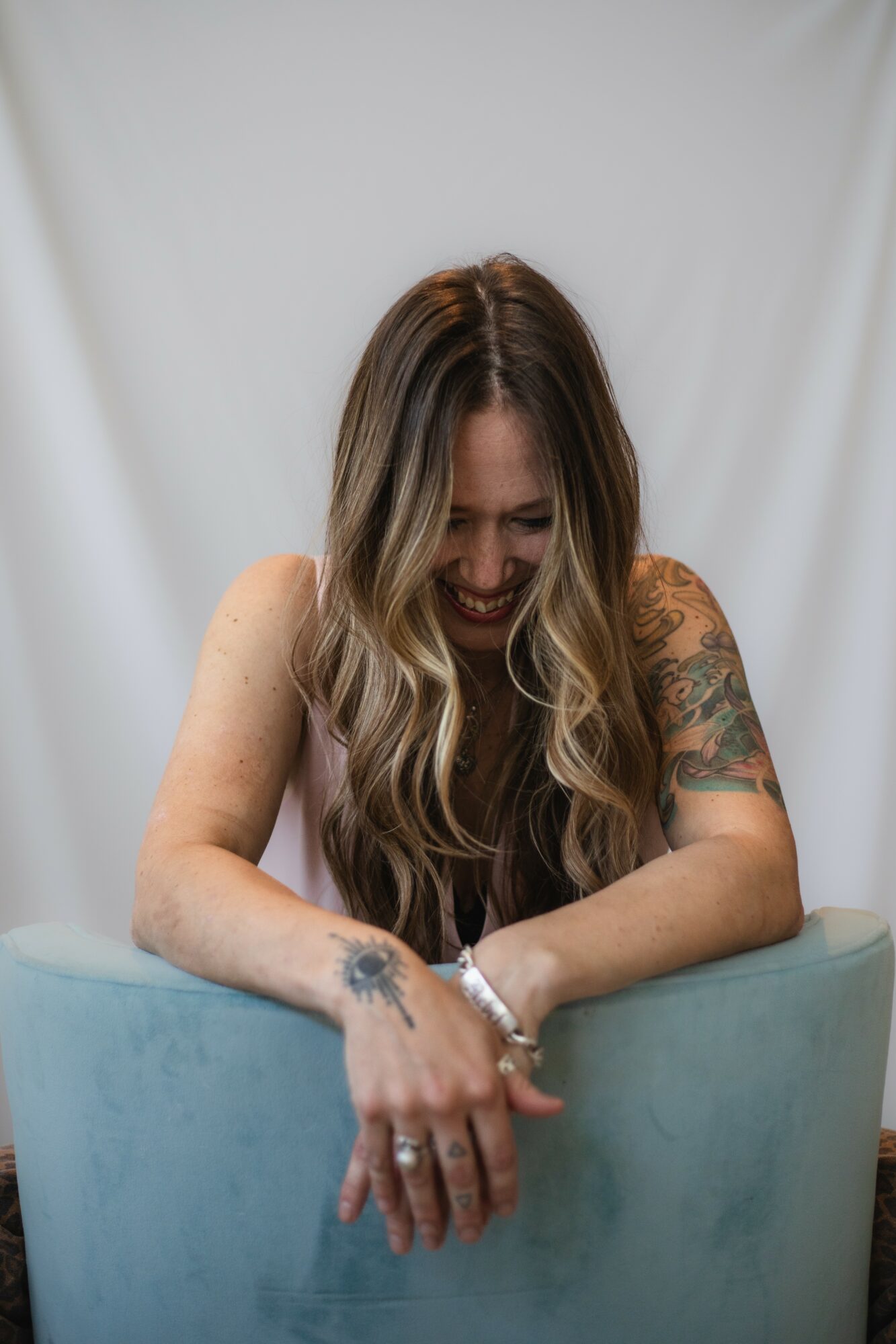 Woman with long wavy hair smiling, leaning on a light blue surface, with a tattooed arm visible.