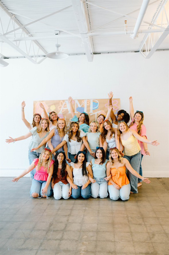 Group of 15 women posing indoors, some raising hands, smiling, with a white wall background.
