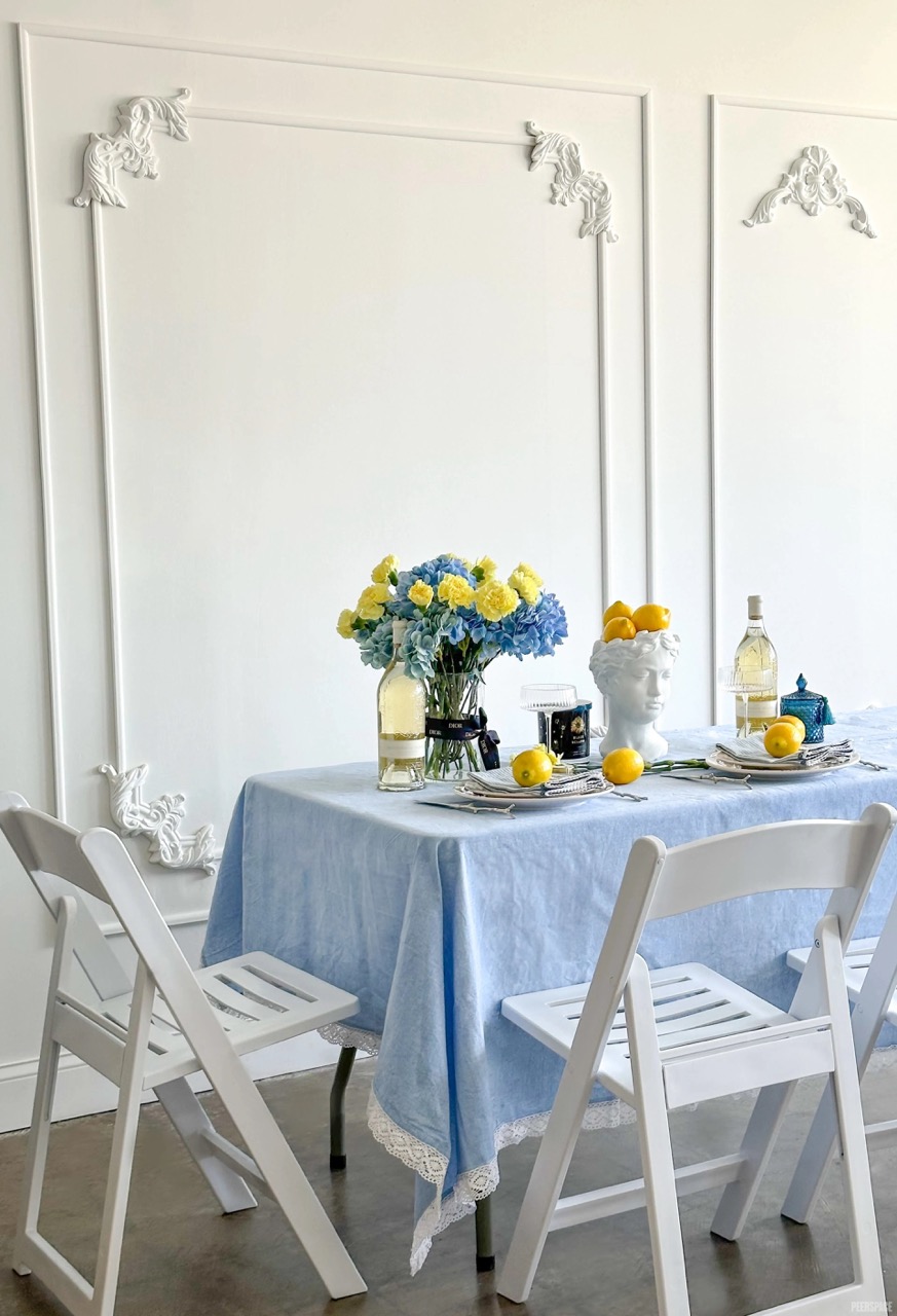 Dining table with blue tablecloth, floral centerpiece, and various bottles and fruits, surrounded by white chairs, against a white wall.