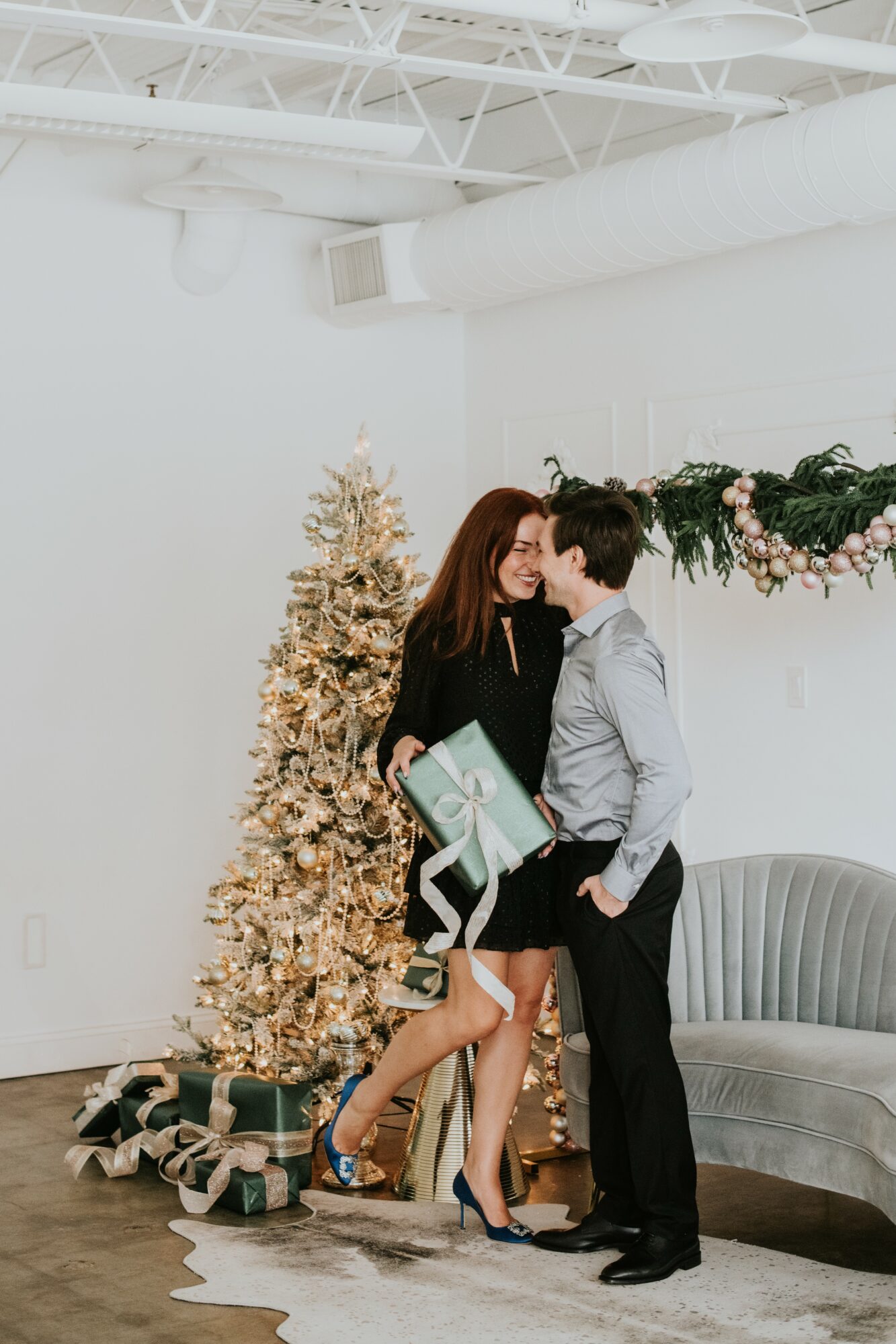 Woman and boy stand close, exchanging gifts near decorated Christmas tree and sofa in bright room.