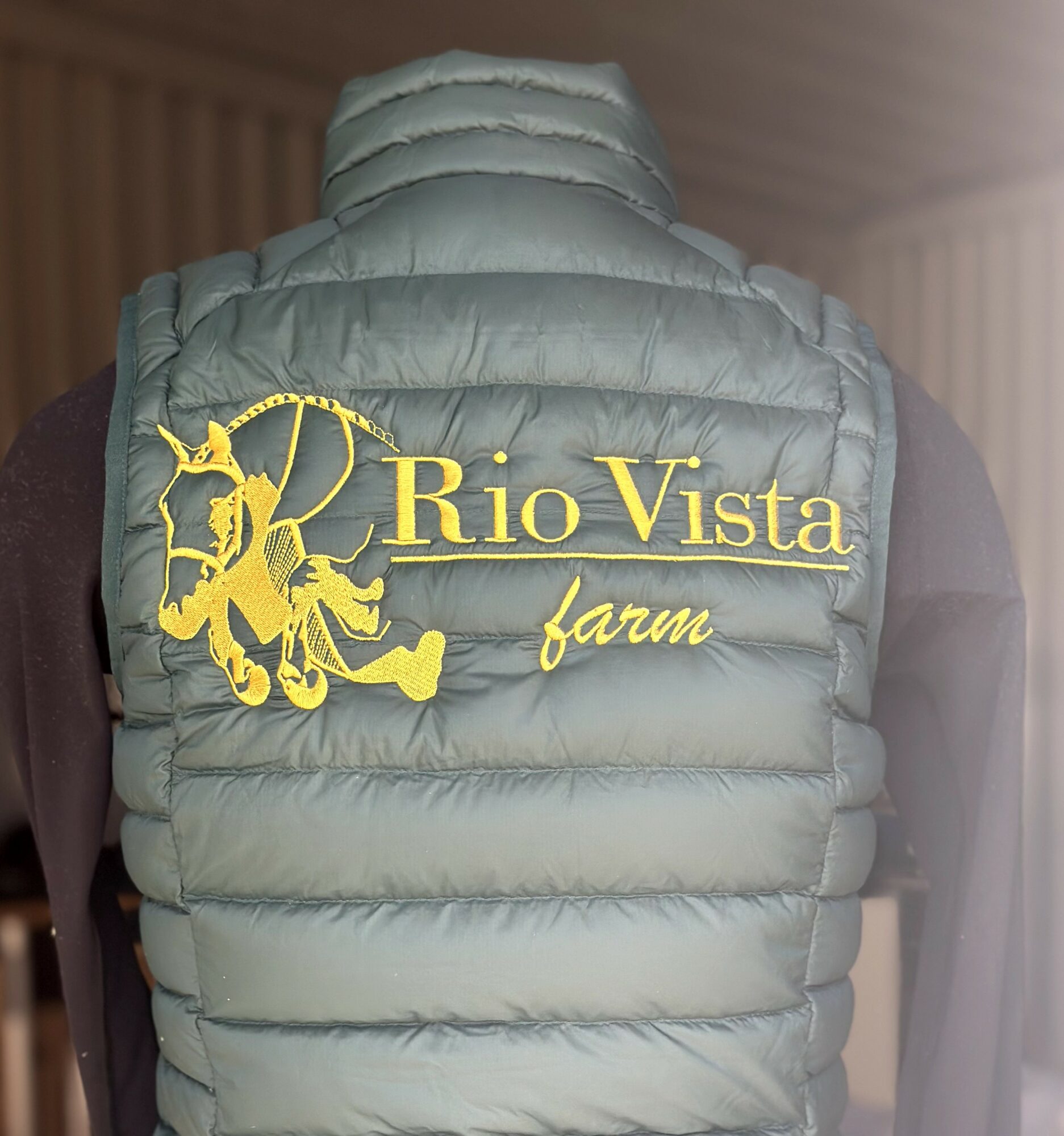 Back view of a light gray puffer vest with yellow embroidery reading 'Rio Vista farm' and a horse logo.