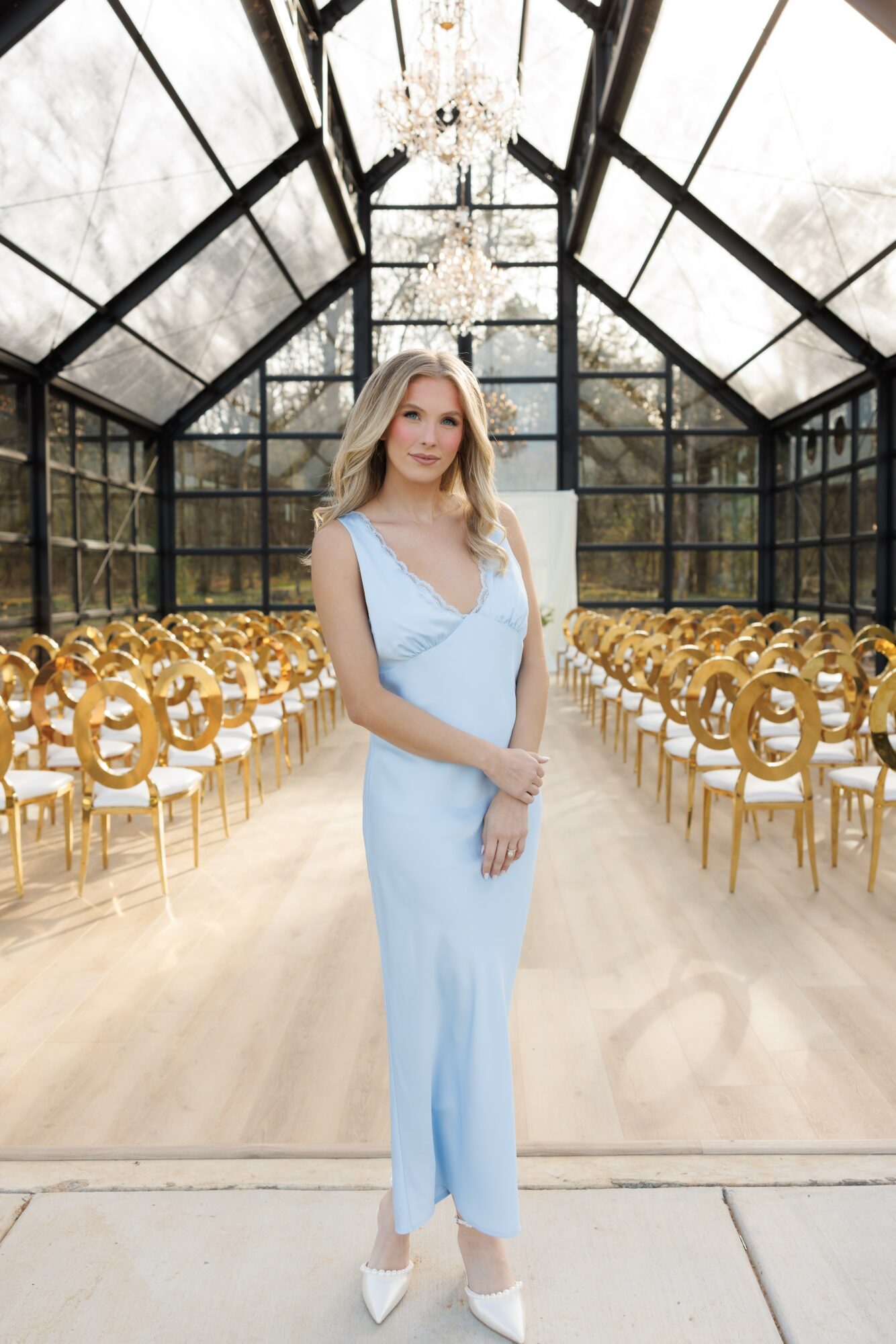 Woman in light blue dress standing in glass-walled venue with gold chairs and chandelier.