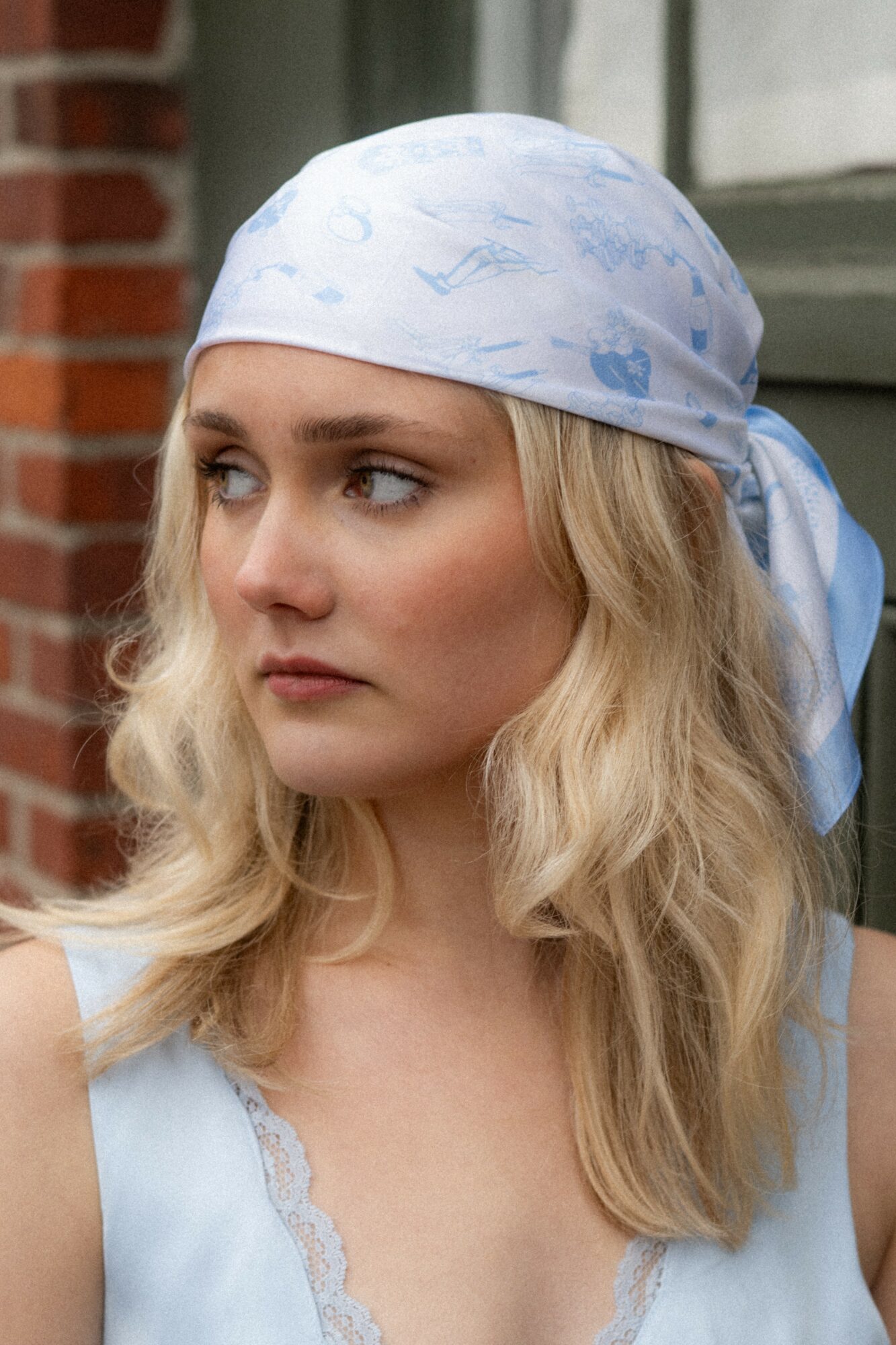Woman with blonde hair wearing a light blue headscarf outdoors, brick wall and window in background.