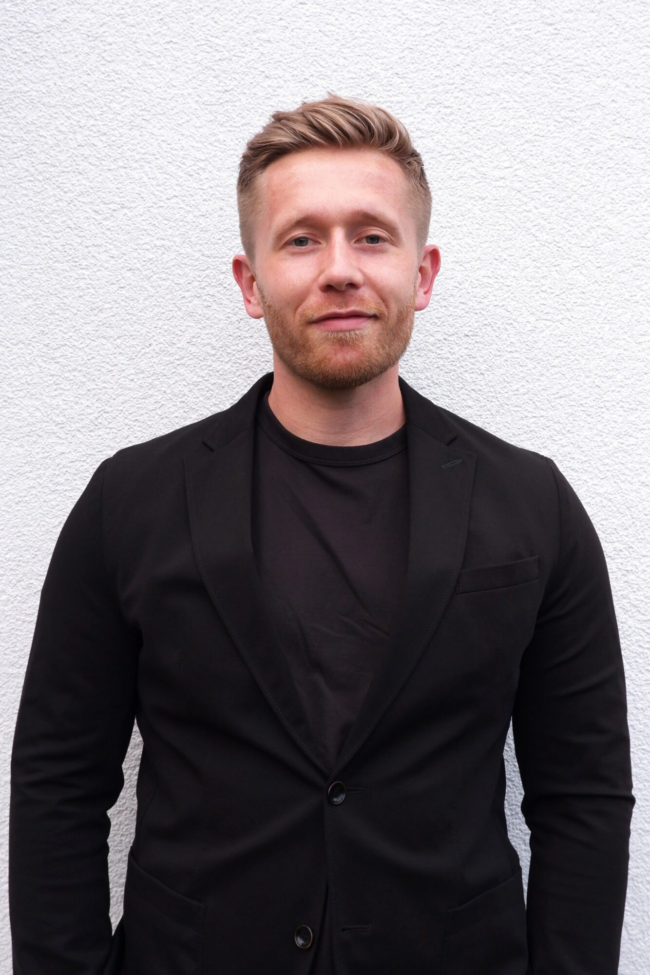 Young man with short blond hair and beard, wearing a black blazer and shirt, standing against a white wall.