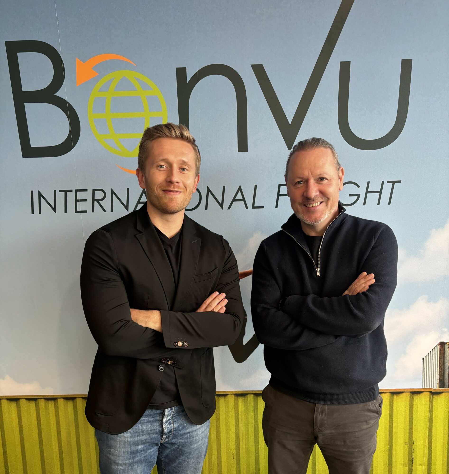 Two men standing side by side with arms crossed, smiling in front of a backdrop with text and logo.