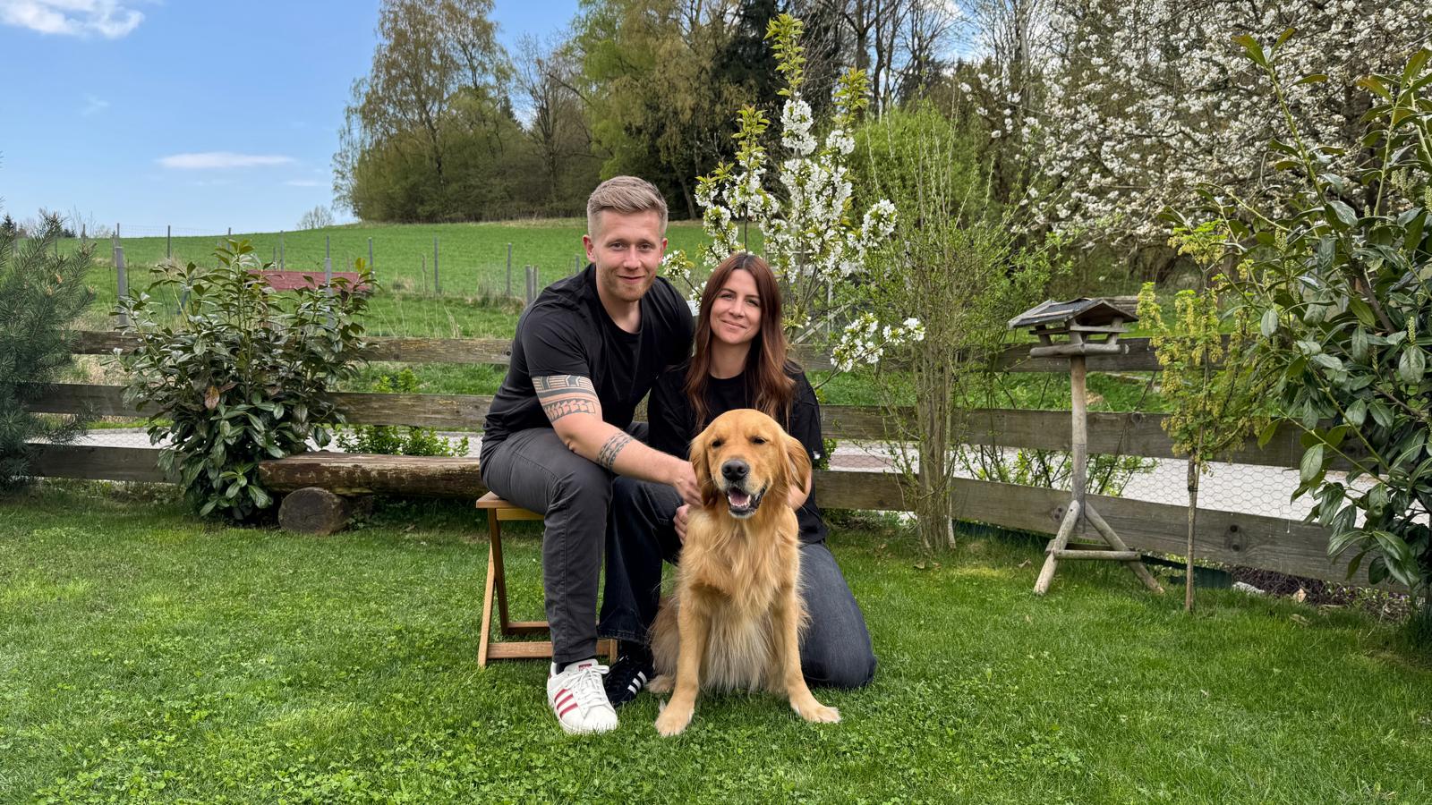 Two people and a dog sitting on grass in a garden with trees and a wooden fence.