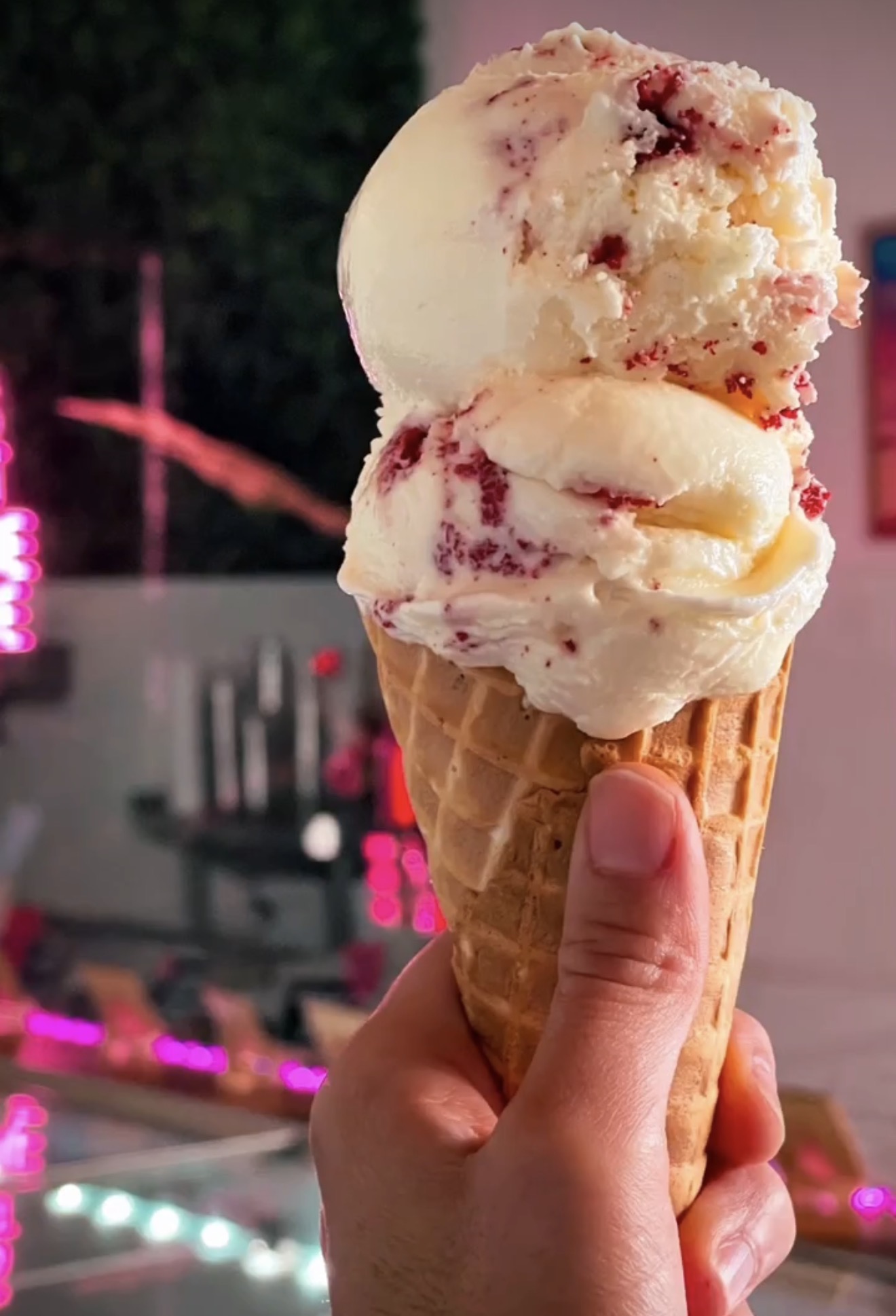 Hand holding a double scoop ice cream cone with pink and white ice cream, outdoors at night.