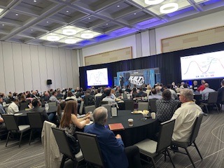 Large conference room filled with people seated at round tables, facing a stage with screens and presentation equipment.