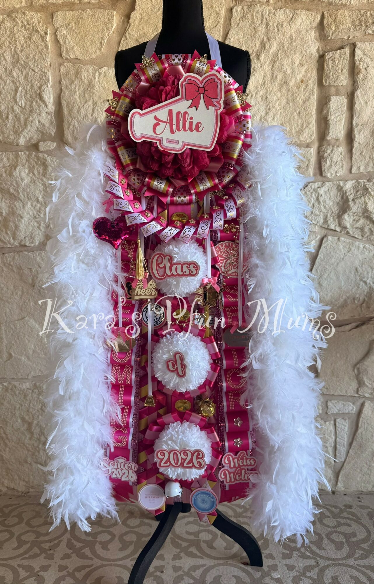 Decorated graduation sash with pink and white ribbons, a heart-shaped tag, and a fluffy white feather boa.