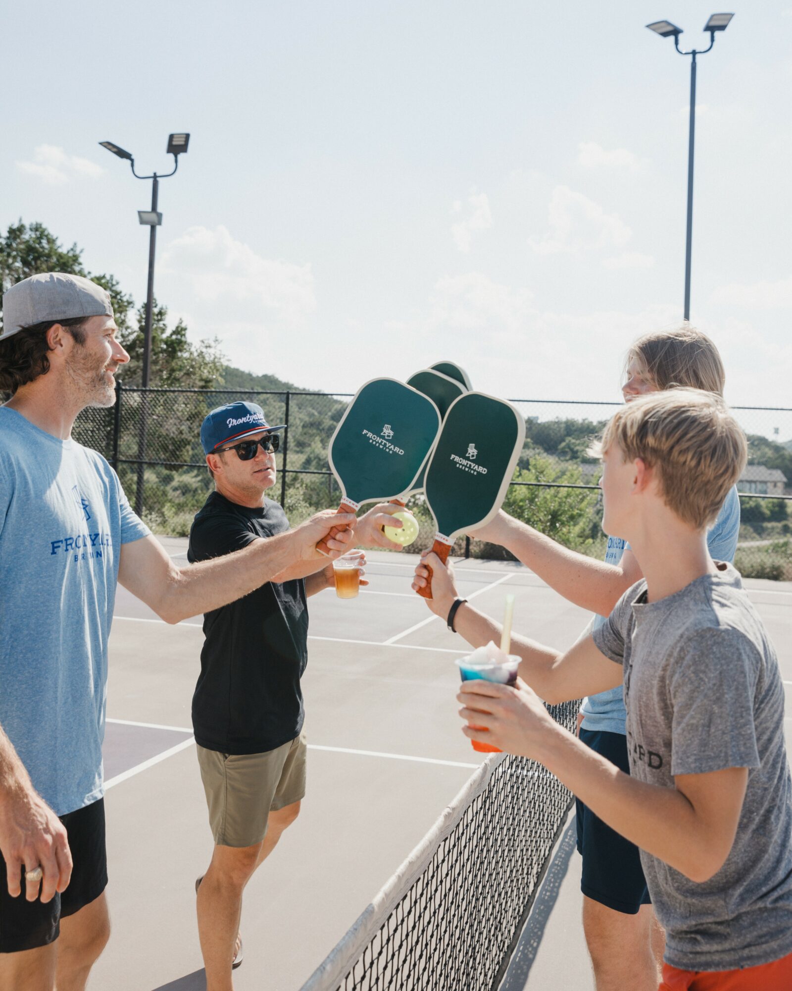 Four people standing outdoors, exchanging drinks over a tennis net, with tennis paddles in the background.