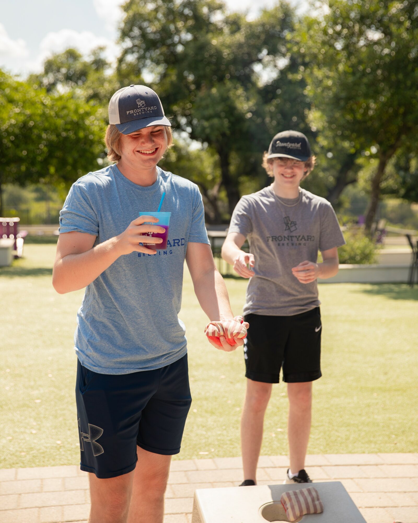 Two boys outdoors, one smiling holding a drink, the other standing on a platform, both wearing caps and casual clothes.