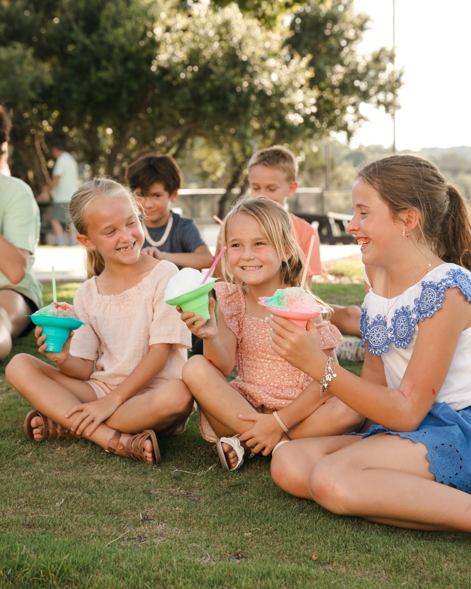 Group of children sitting on grass, smiling, holding colorful bowls, outdoors with trees in background.