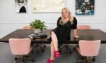 Woman with long blonde hair in black dress sitting on conference table with pink chairs, artwork on wall, plant, and books.