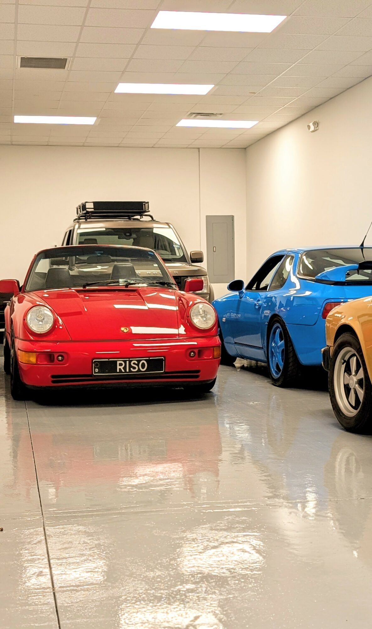 Multiple sports cars parked indoors, including a red car with a license plate reading RISO, and a blue car beside it.