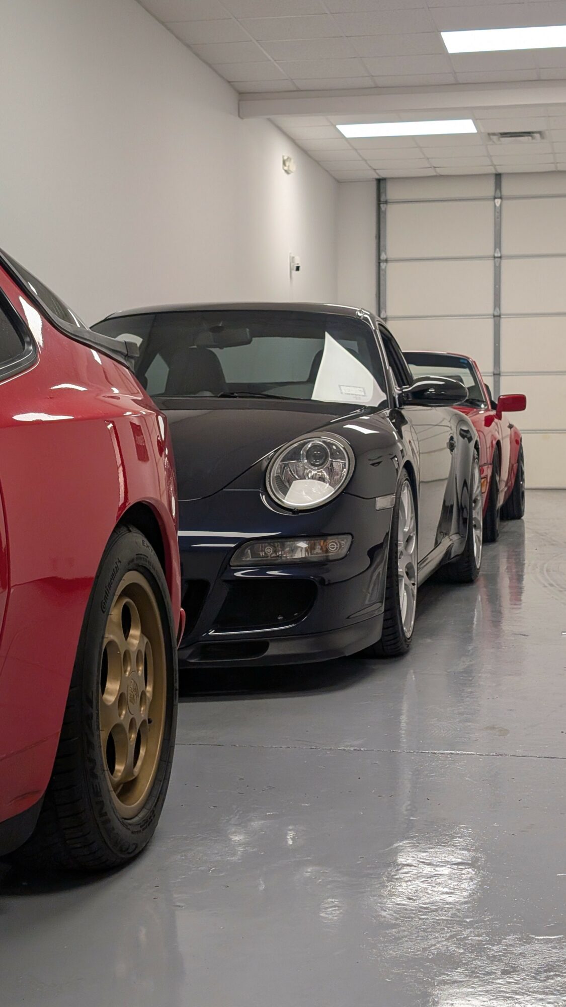 Black sports car parked indoors with a red vehicle partially visible in the foreground.