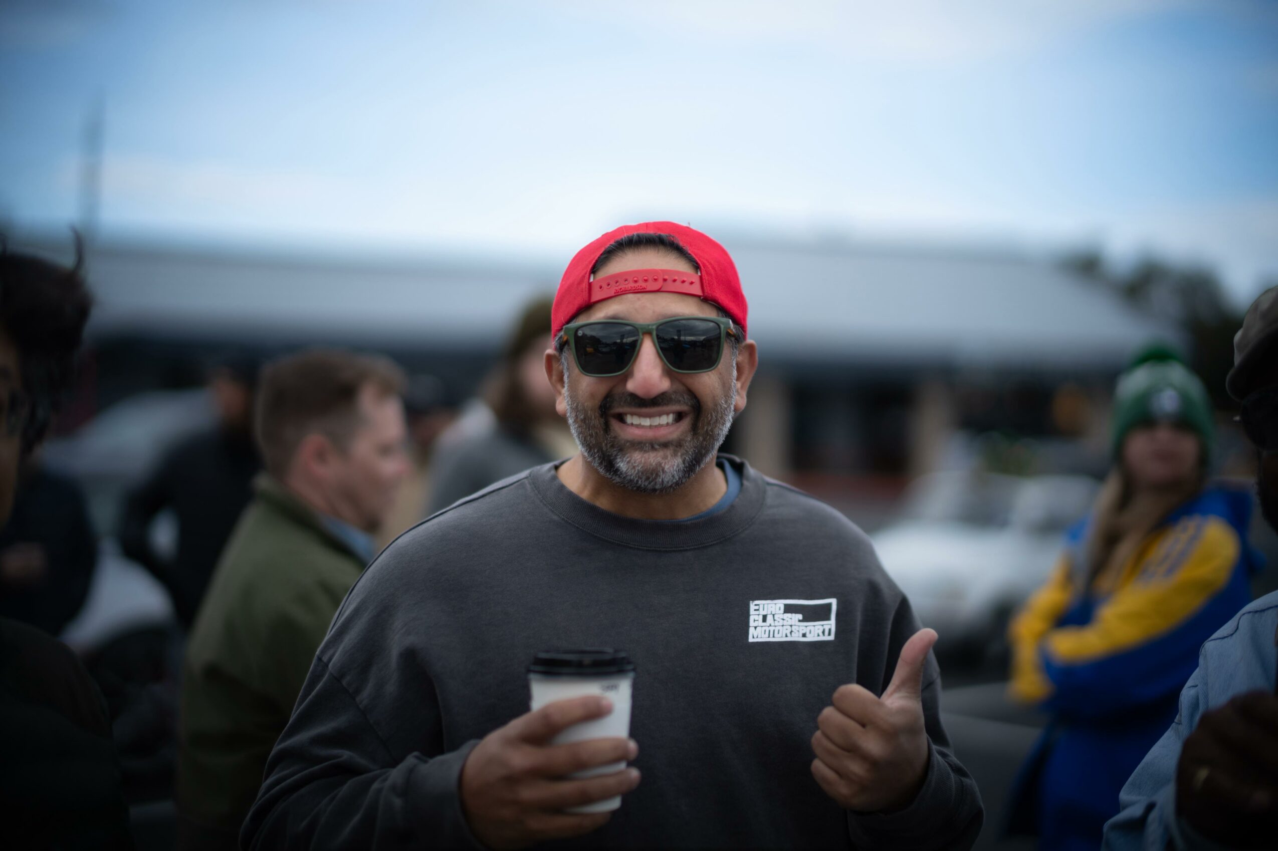 Man wearing sunglasses and a red cap smiling and giving a thumbs-up at an outdoor event.