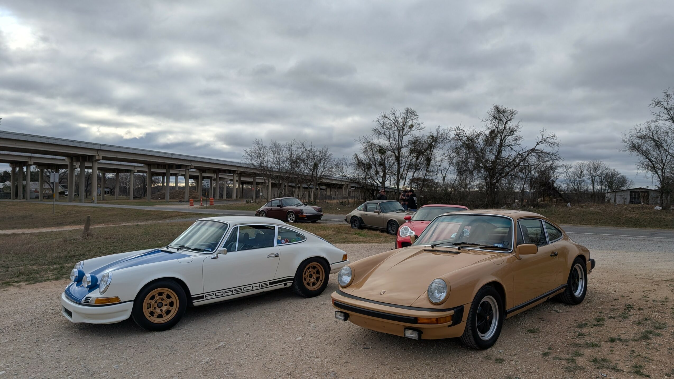 Two classic cars parked outdoors on a cloudy day, with trees and a building in the background.