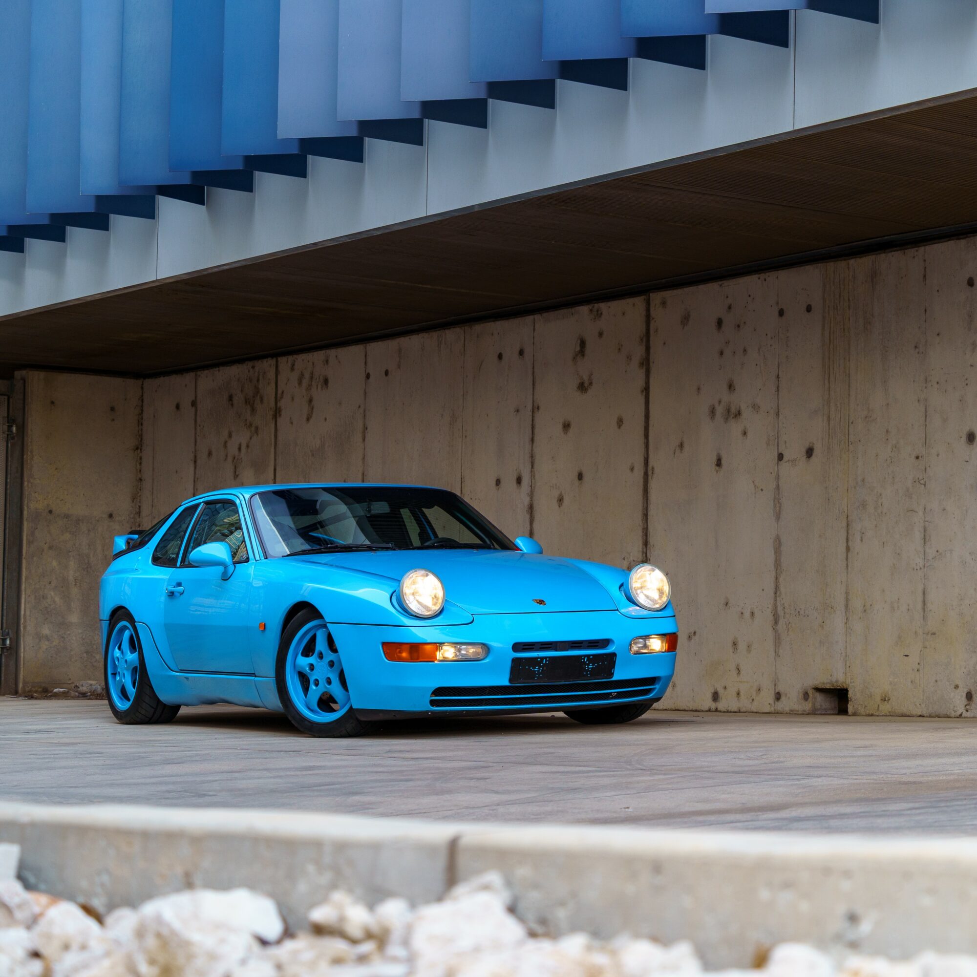 Blue sports car parked against a concrete wall under a modern building with blue and white architectural elements.
